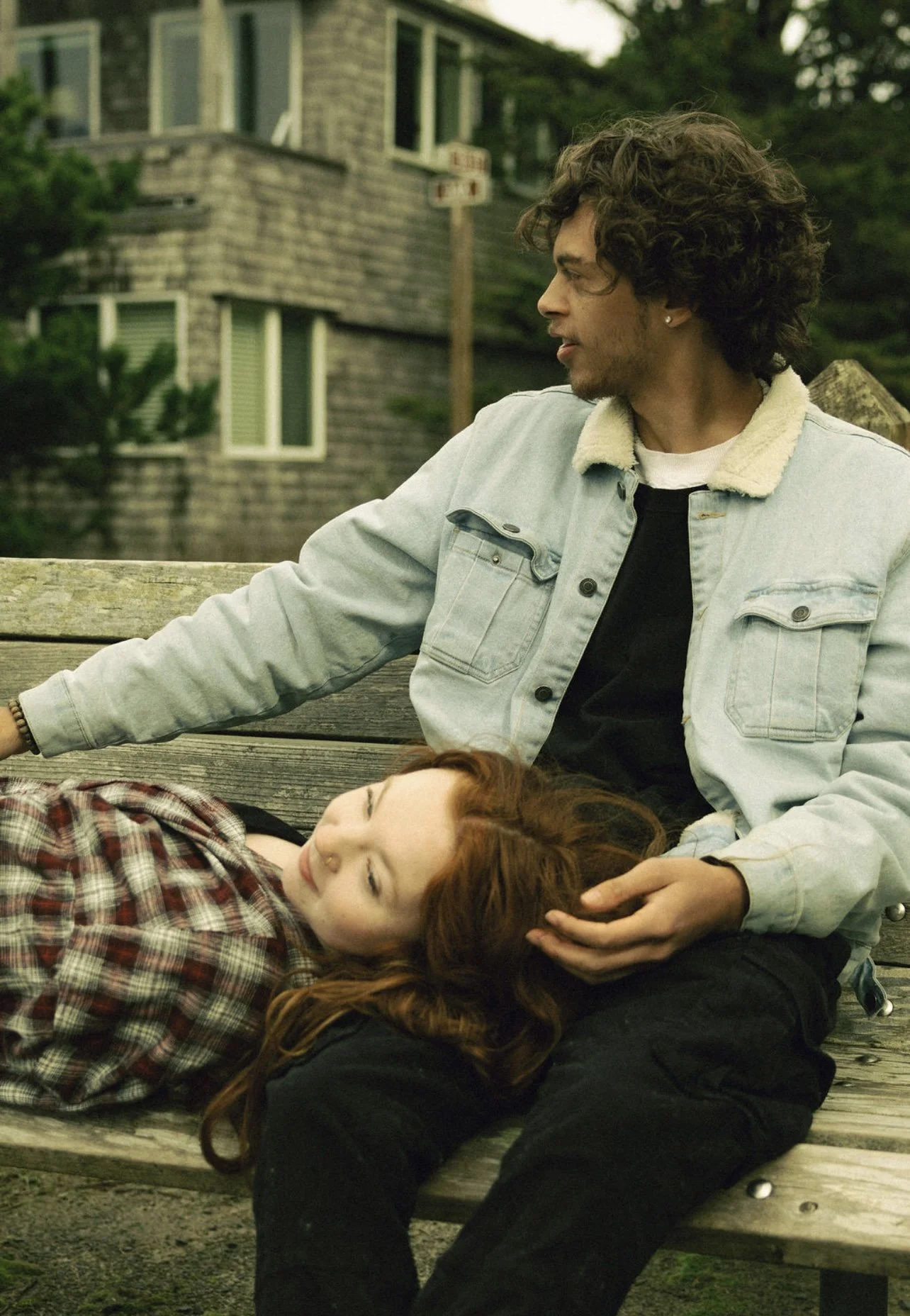 A young man with curly hair and earrings sitting on a wooden bench with a young woman with red hair laying down with her head on his lap. They are outdoors in front of a house with a shingled exterior.