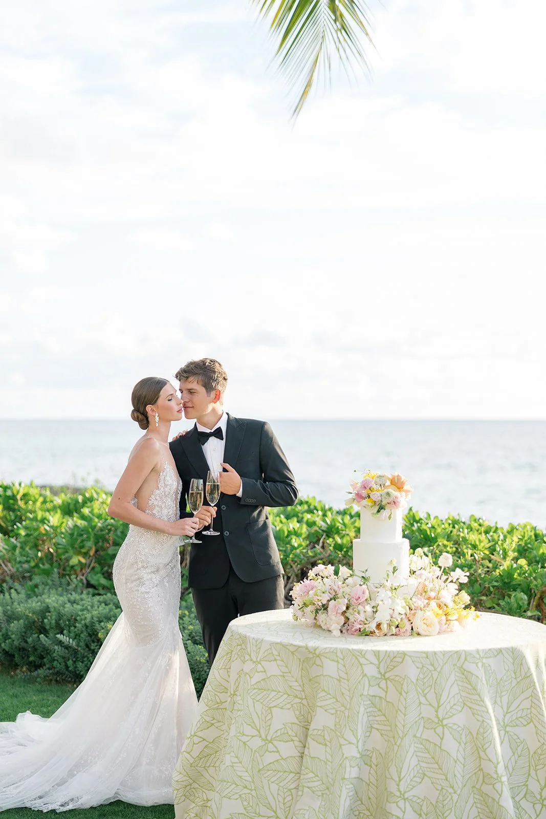 A bride and groom in wedding attire holding champagne glasses, standing near a table with a white wedding cake decorated with flowers, on a grassy area near the ocean, with a palm tree leaf visible in the top right corner.