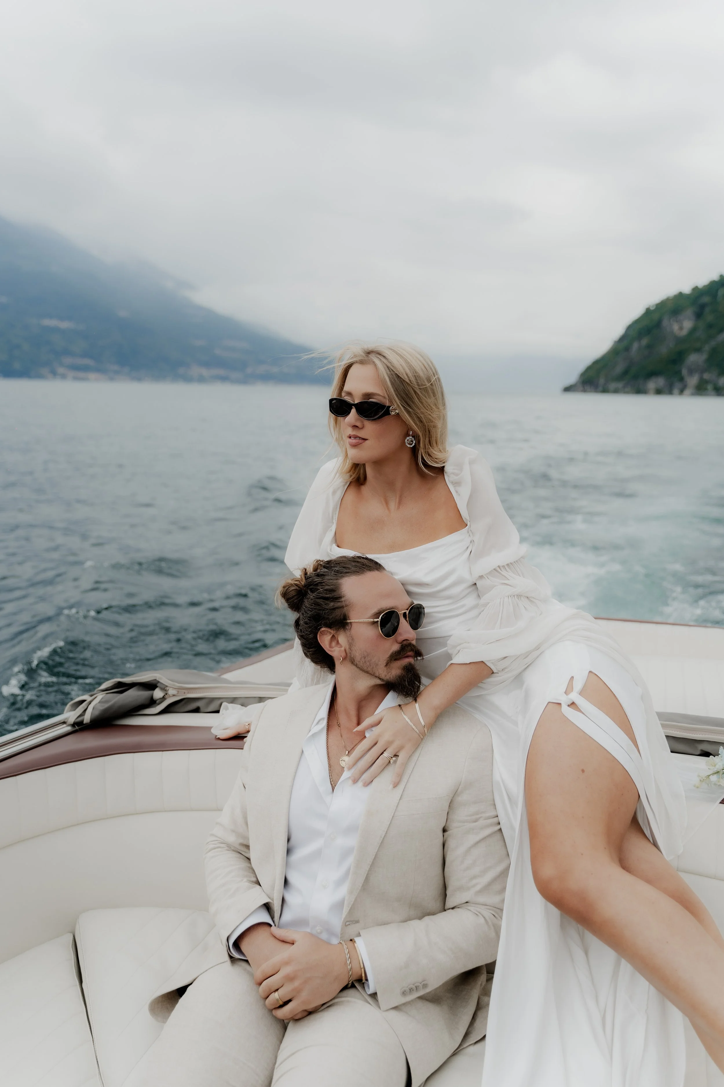 A stylish woman and a man sit on a boat with a scenic water and mountain backdrop, both wearing sunglasses and dressed in white, with the woman resting her hand on the man's shoulder.