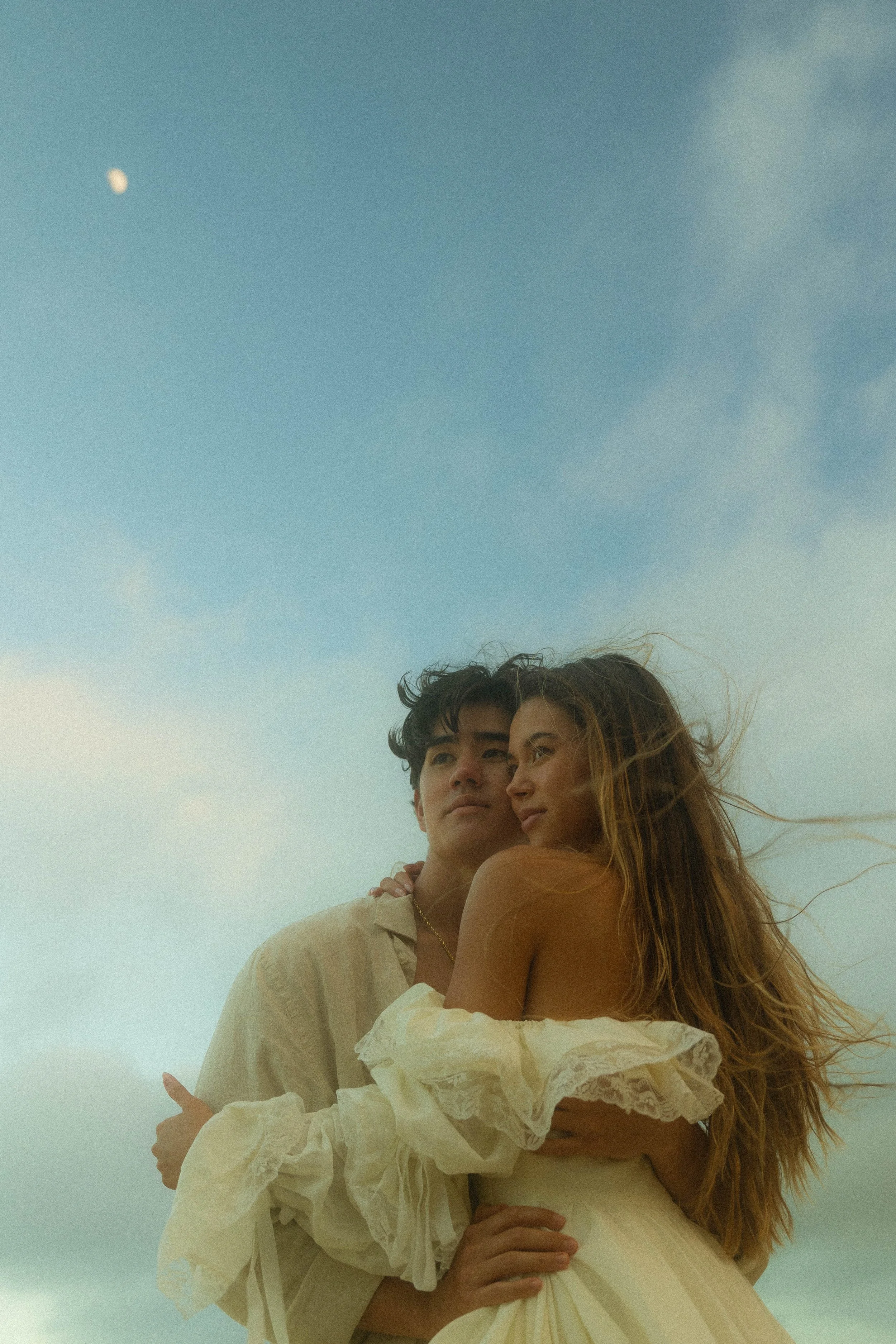 A young man and woman embrace outdoors under a partly cloudy sky with the moon visible in the distance.