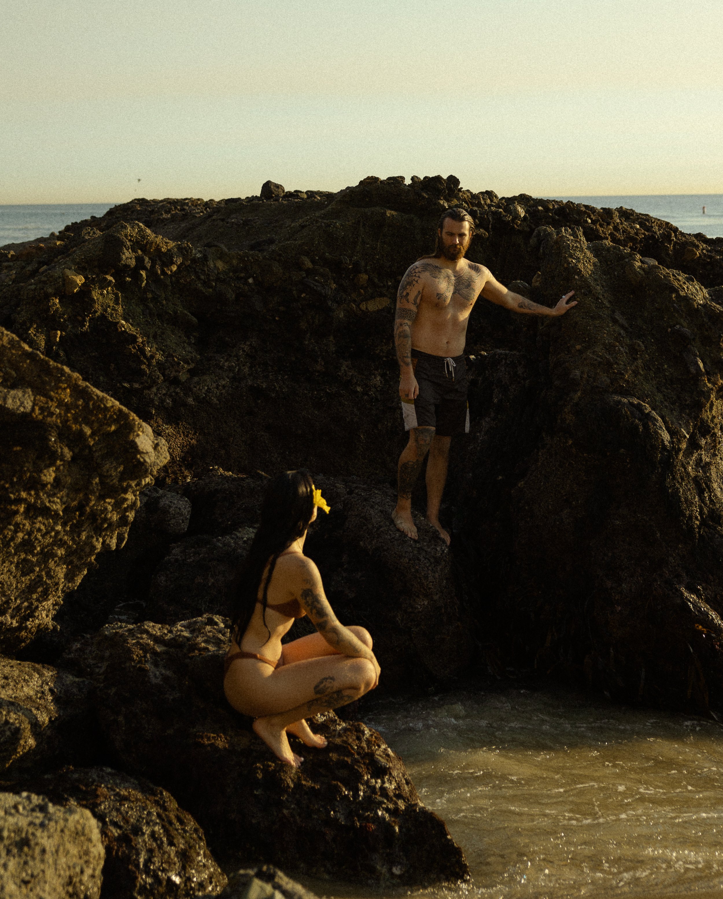 A bearded man with tattoos standing on rocks near the water, and a woman with long dark hair and tattoos sitting on rocks closer to the water, both at a beach during sunset.