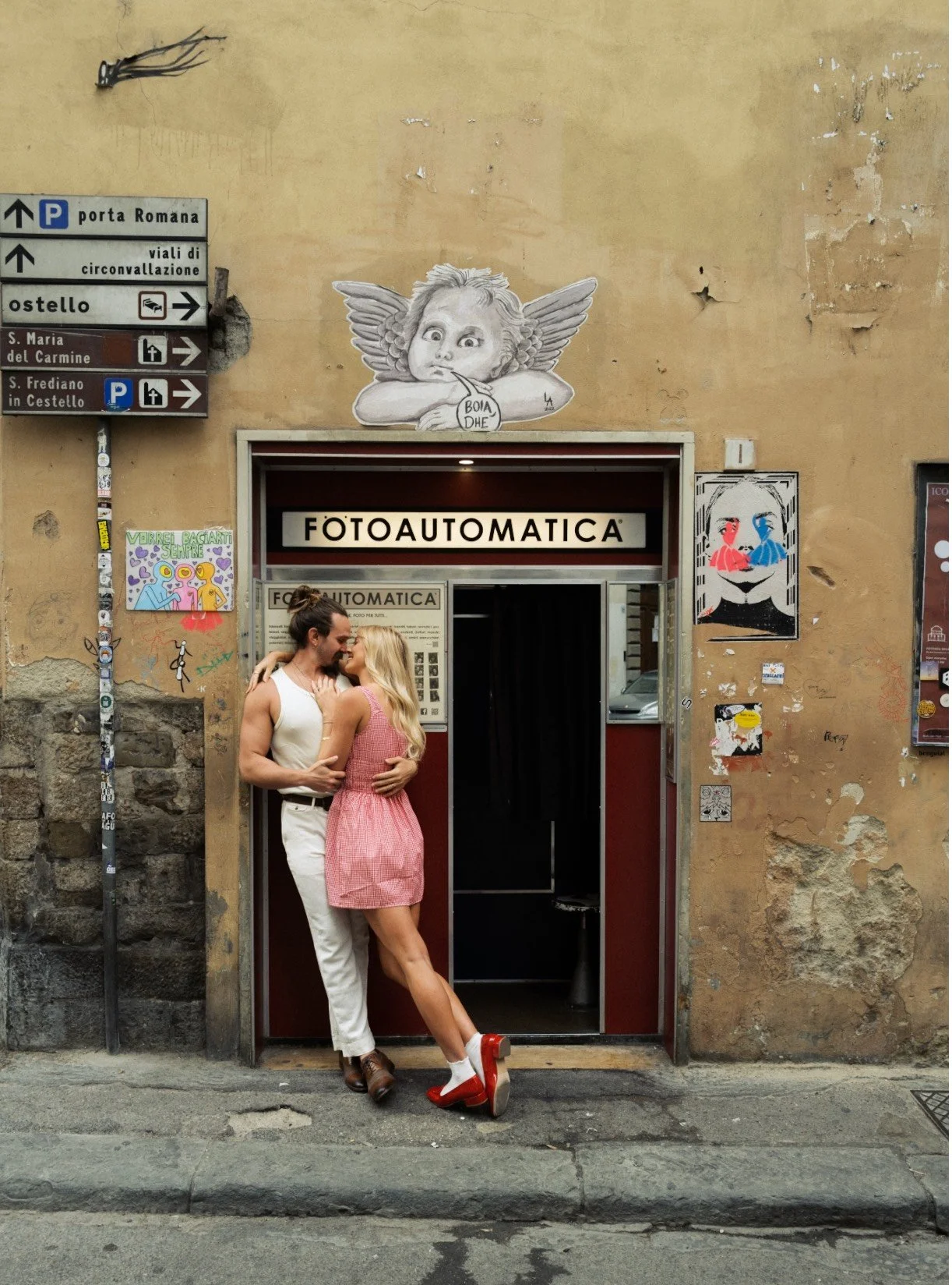 A couple kissing outside a photo booth beneath street signs and graffiti art on a weathered wall.