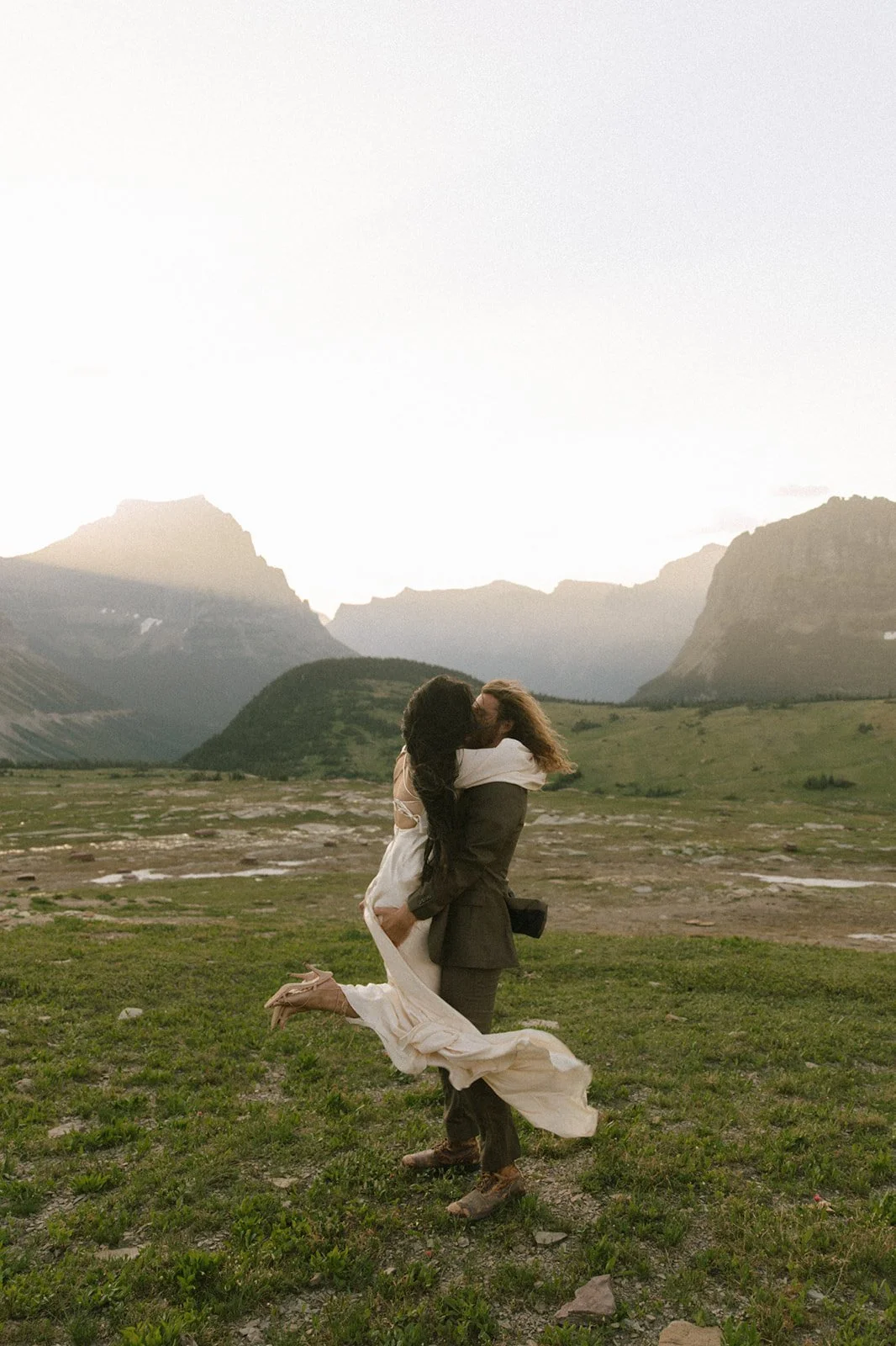 A couple kissing in a scenic mountainous landscape, with the woman wearing a white dress and the man in a suit, as the man lifts the woman off the ground.
