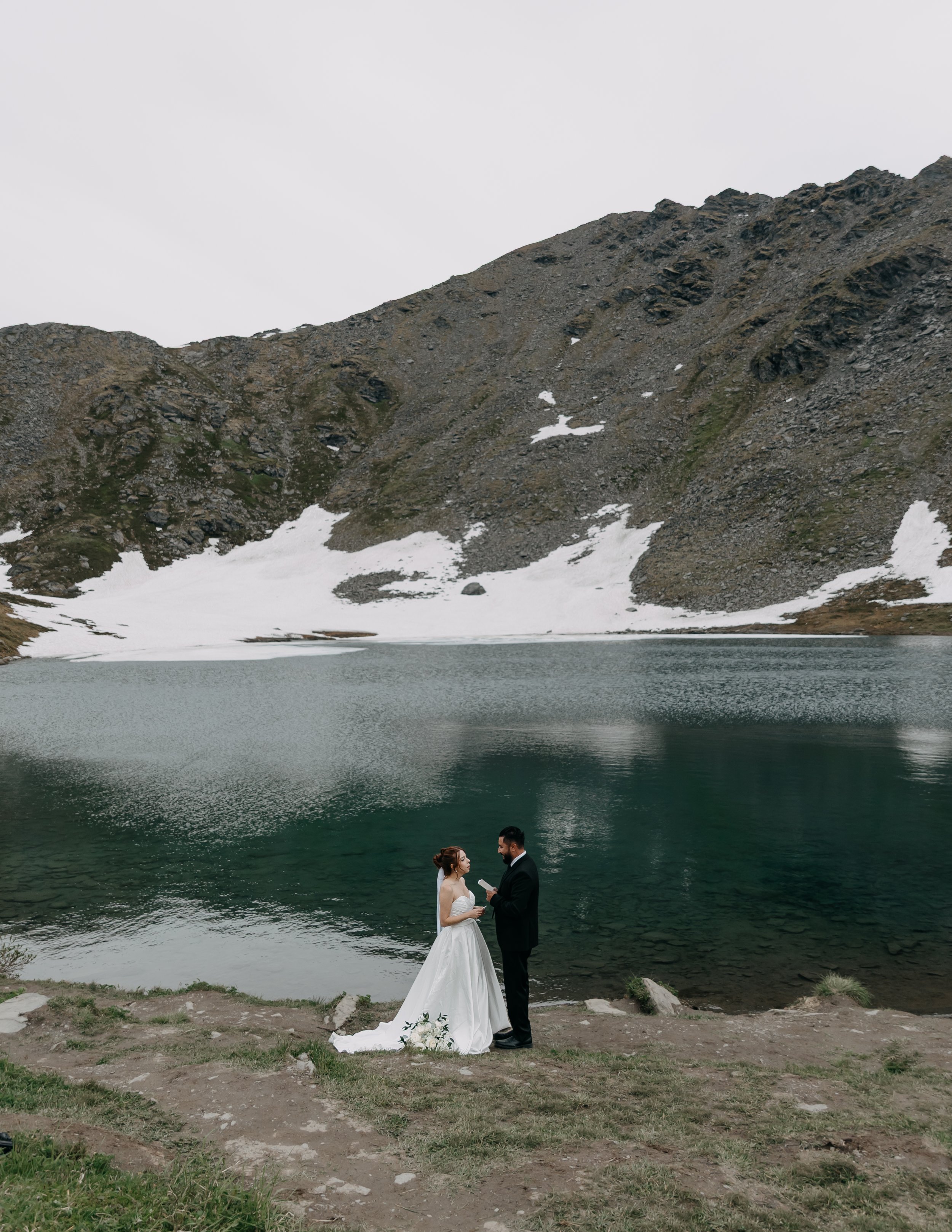 A bride and groom in wedding attire standing by a mountain lake, exchanging vows in an outdoor setting with snow patches on the mountainside.