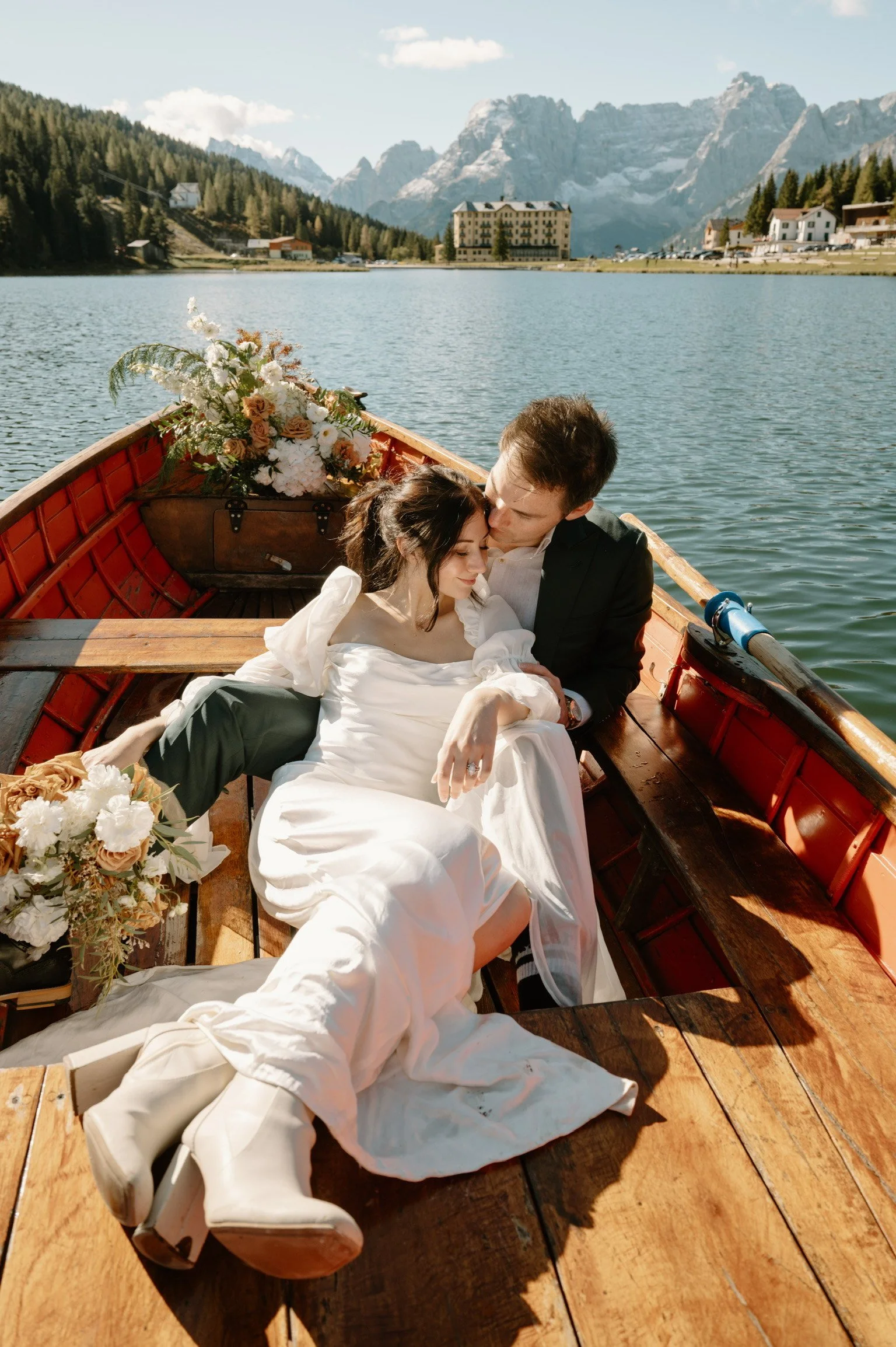A newlywed couple sits closely in a wooden boat on a lake, surrounded by mountains, with a bouquet of flowers nearby and a backdrop of a mountain village.