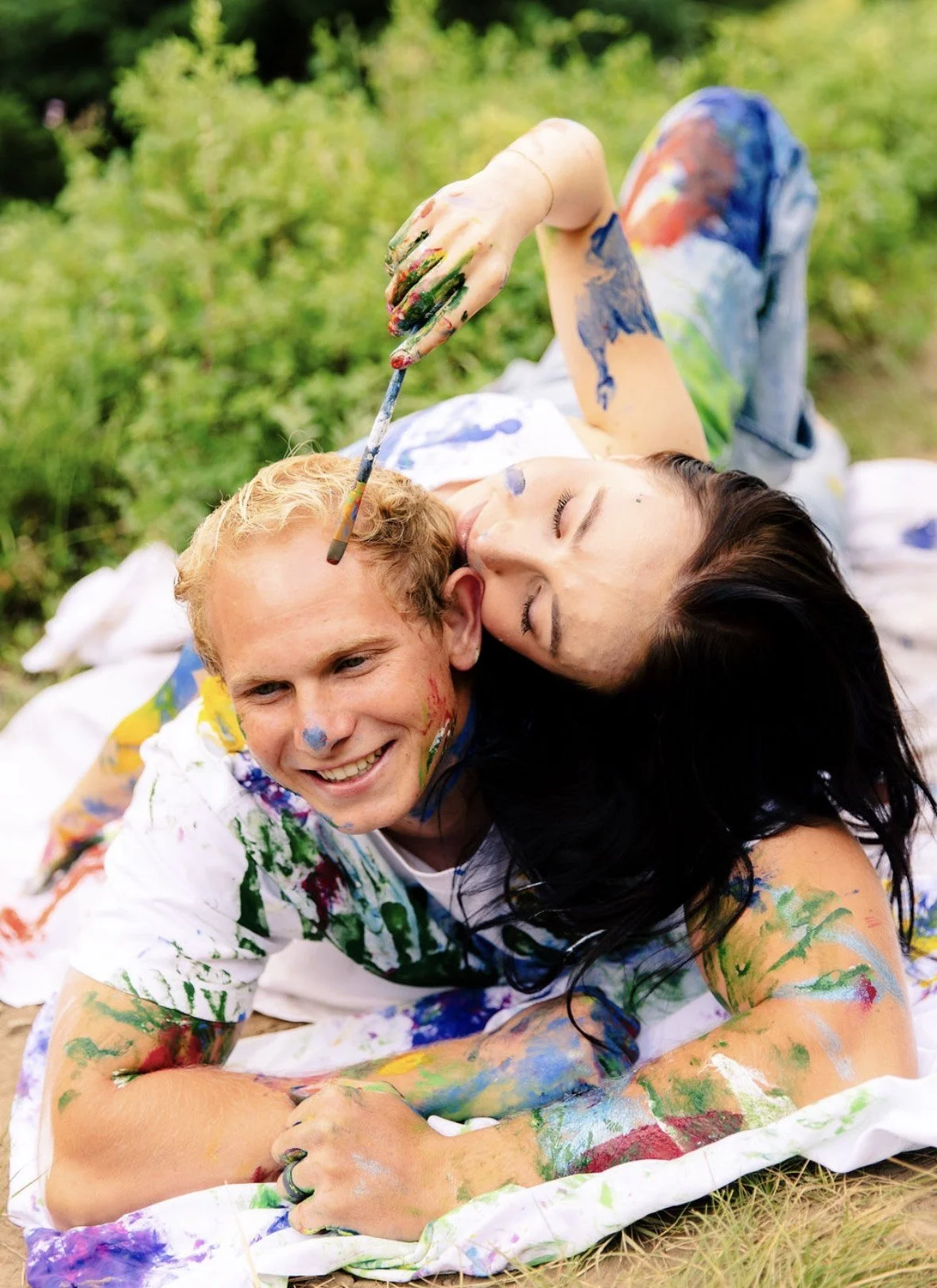 Two people lying on the grass covered in colorful paint, smiling and playing with paintbrushes.