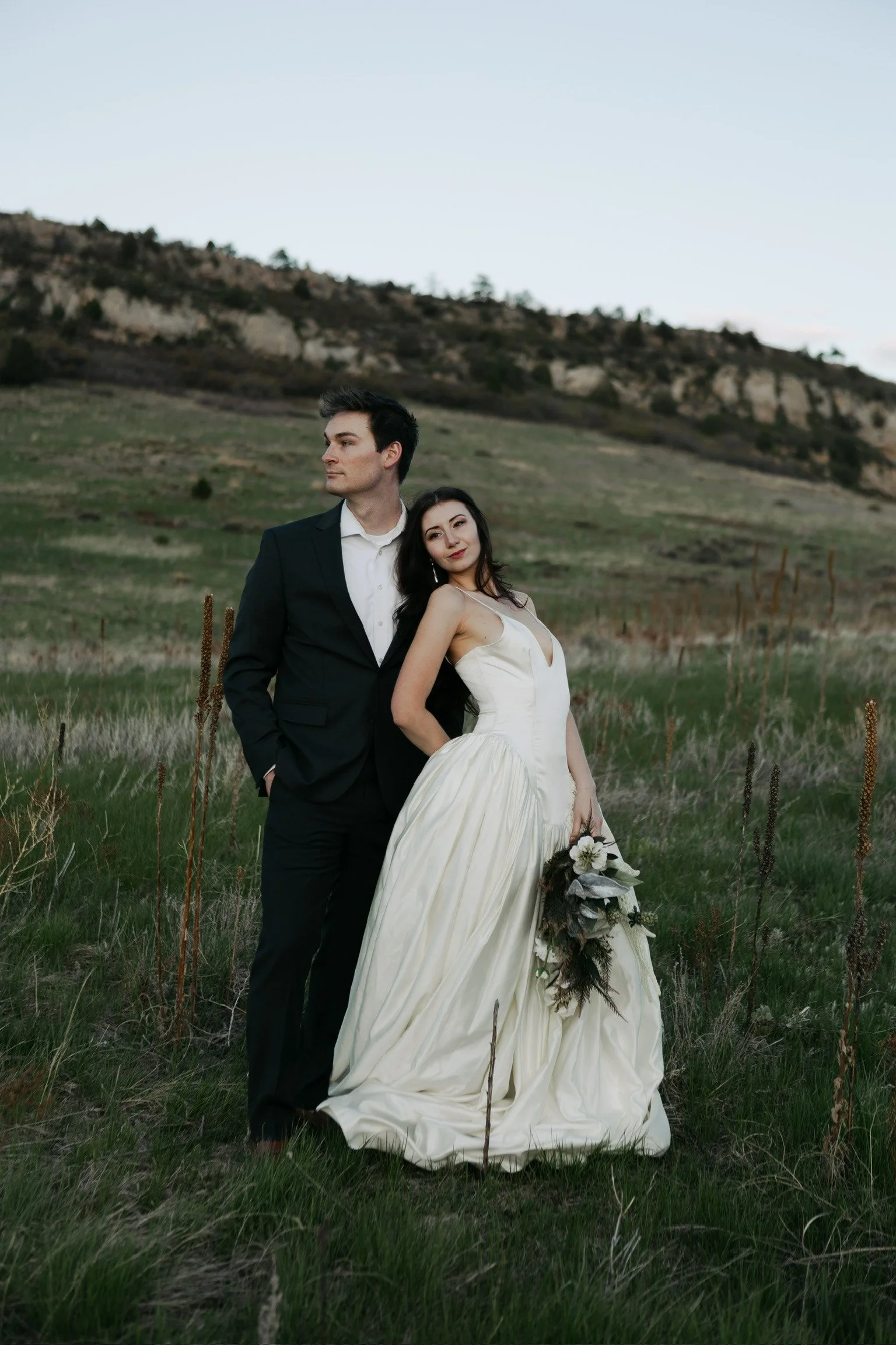 A man in a black suit and a woman in a white wedding gown standing in a grassy field with hills in the background, holding a bouquet.