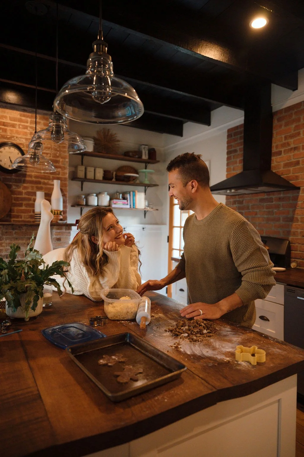 A man and a woman smiling at each other in a cozy kitchen, preparing cookies together. The woman is sitting at a wooden kitchen island, and the man is standing next to her, leaning on the counter. There are baking utensils and ingredients on the counter, and the kitchen has a warm, rustic decor with brick walls and open shelving.