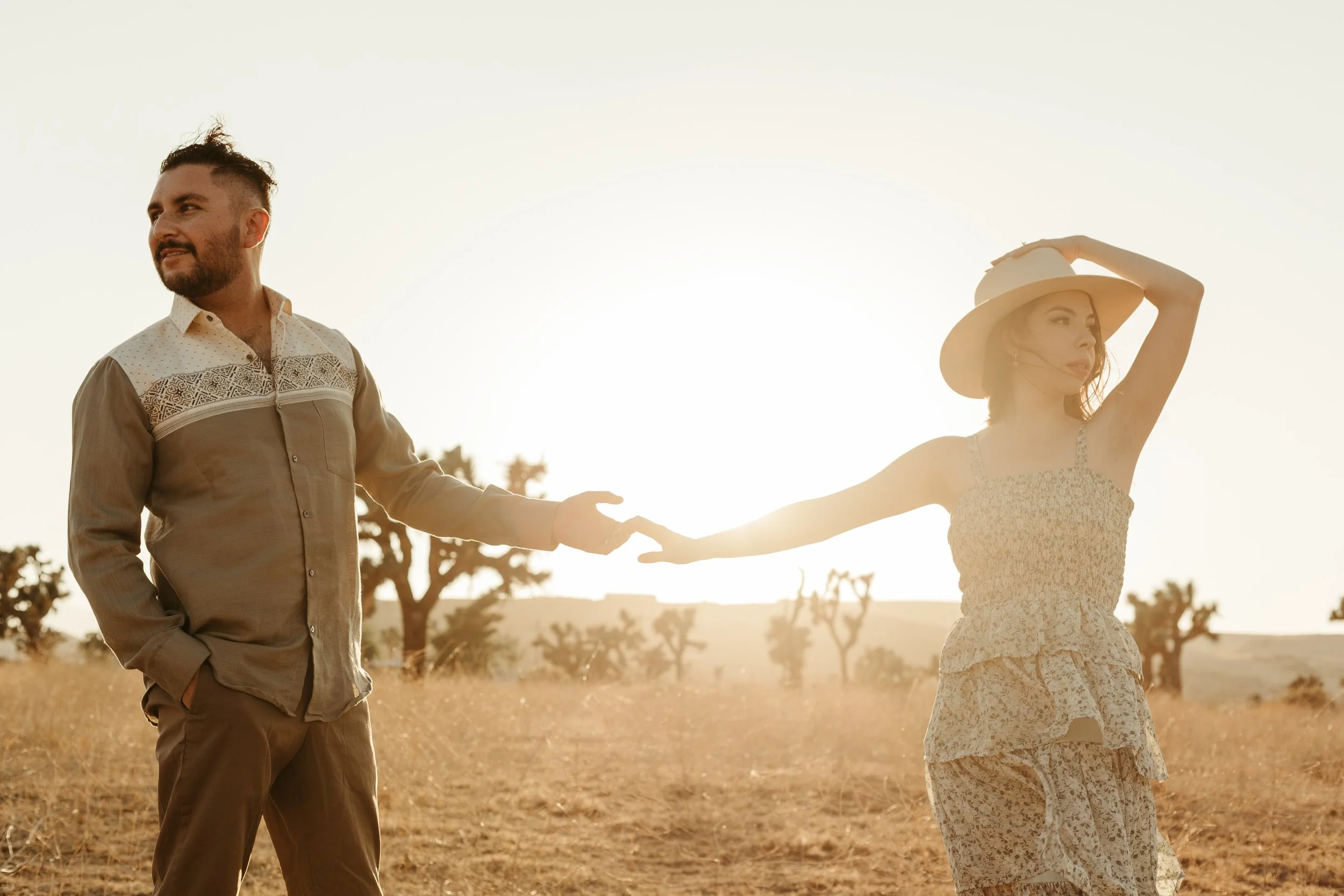 A man and woman holding hands in a desert landscape during sunset. The woman wears a floral dress and wide-brimmed hat; the man wears a patterned shirt and pants.