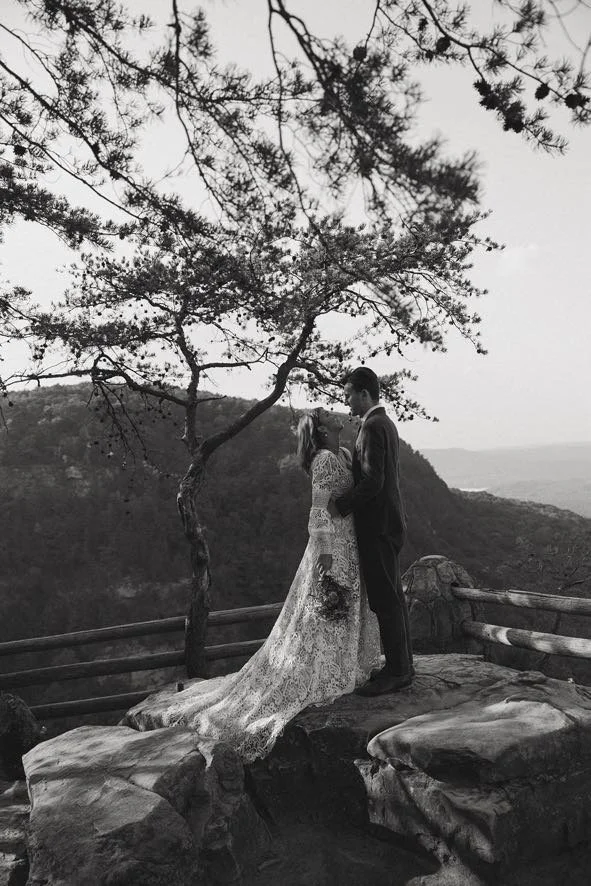A black-and-white photo of a couple, dressed in wedding attire, standing on a rocky ledge near a tree with mountains in the background.