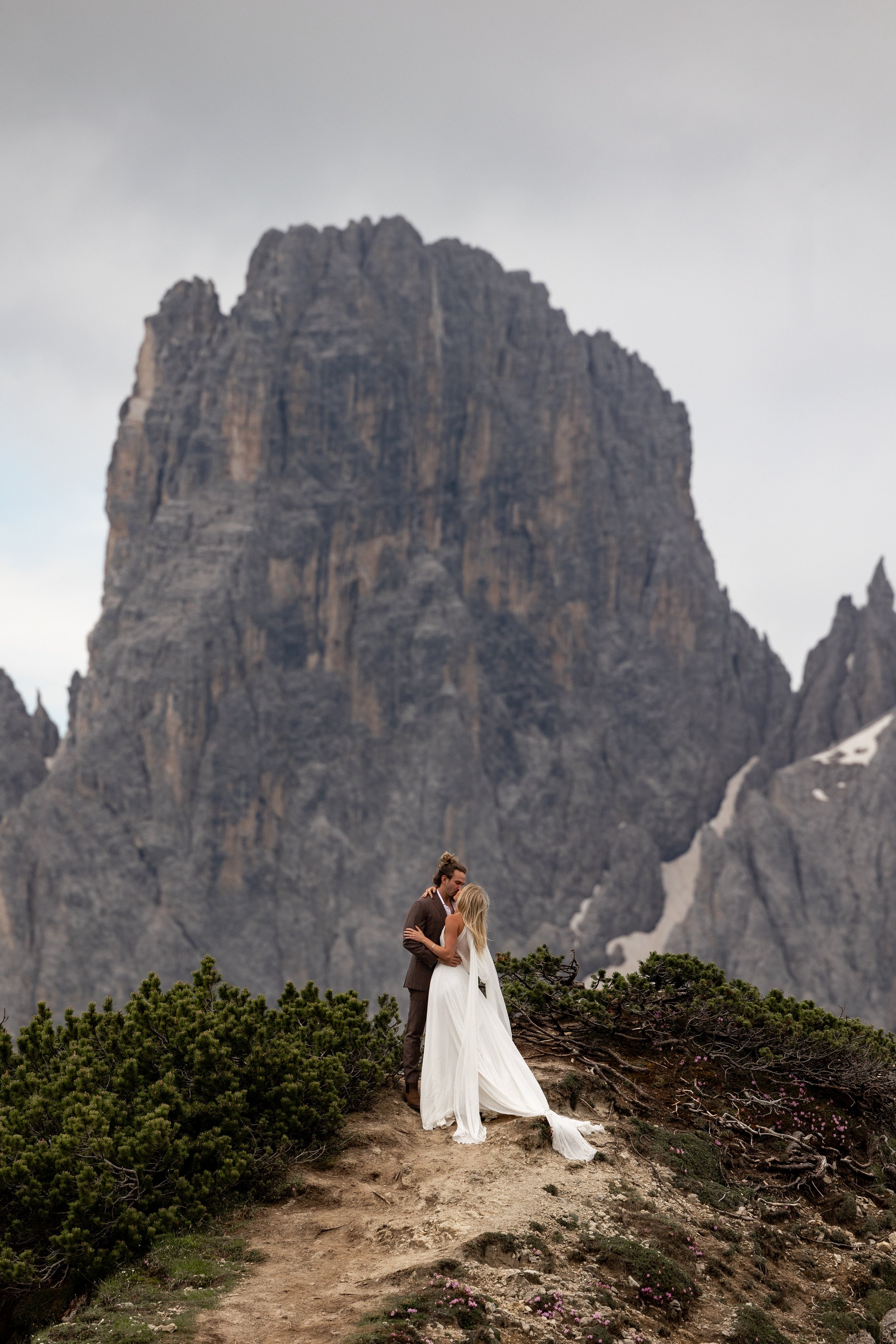 A couple dancing on a mountain trail with a large, rugged mountain in the background during cloudy weather.