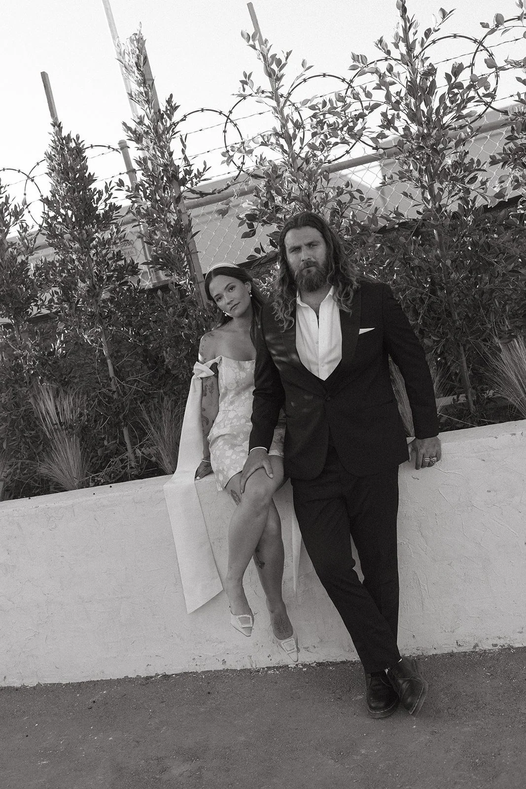 A woman in an off-shoulder dress with floral pattern sitting on a ledge, and a man in a suit leaning against the same ledge in front of a fence with barbed wire and plants, black-and-white photo.