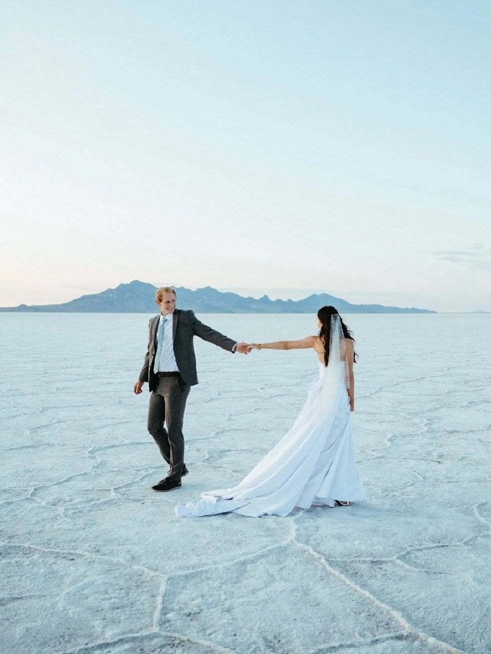 A bride and groom holding hands on a salt flat, with mountains in the background during sunset.