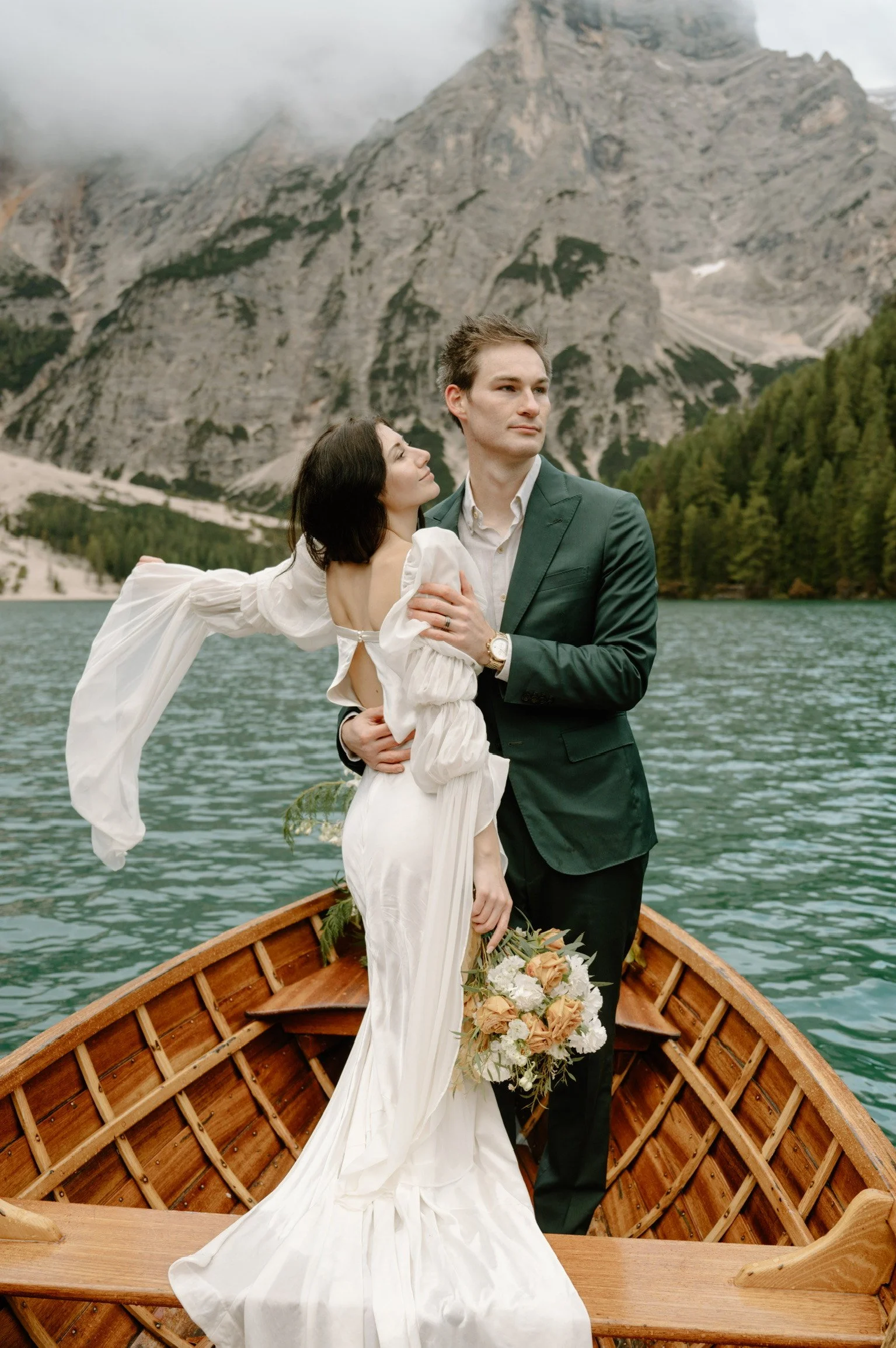 A bride and groom standing in a wooden boat on a lake with mountains and pine trees in the background. The bride is holding a bouquet of flowers, and the groom is wearing a dark green suit.