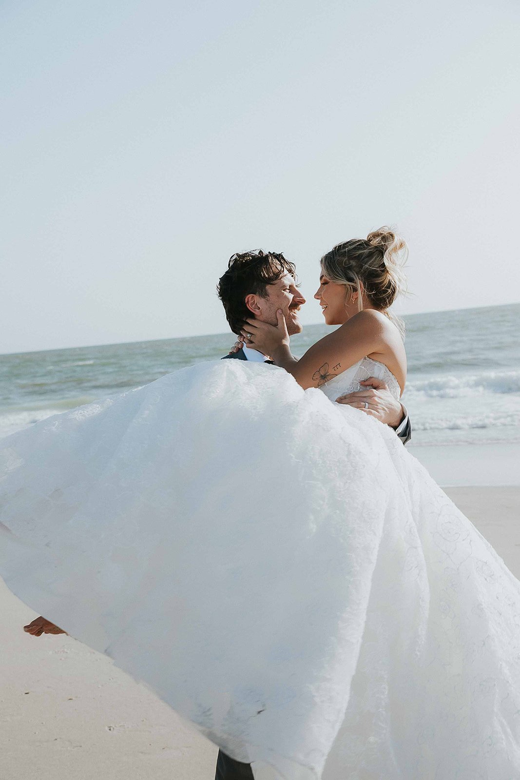 A couple in wedding attire on the beach, smiling and embracing with the ocean in the background.