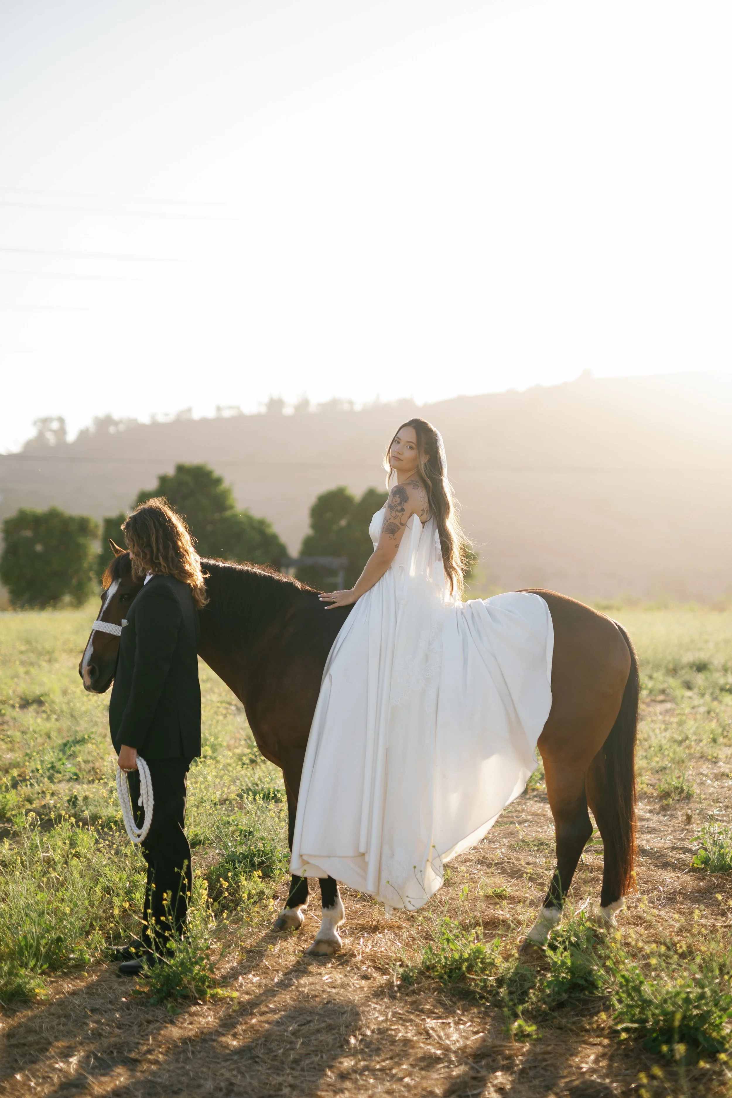 A woman in a white wedding dress sitting on a brown horse during sunset in an open field, with a man in a black suit holding the horse's reins.