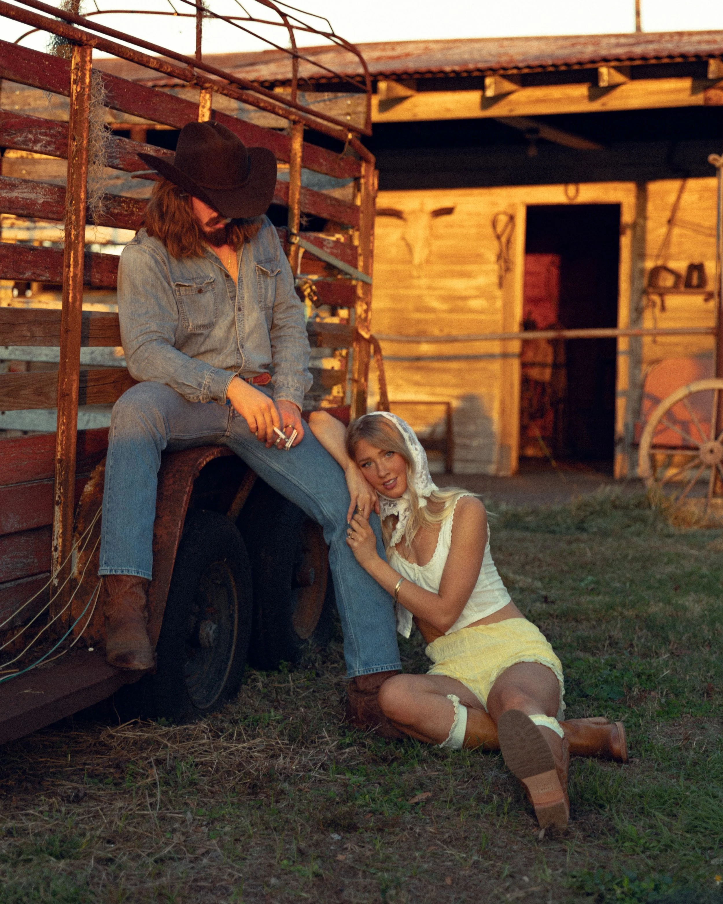 A man wearing a cowboy hat, denim shirt, and jeans sitting on a rusty wagon. A woman in a yellow skirt, white top, and brown boots kneels on the ground, holding his leg and leaning on it, in front of a rustic wooden barn.