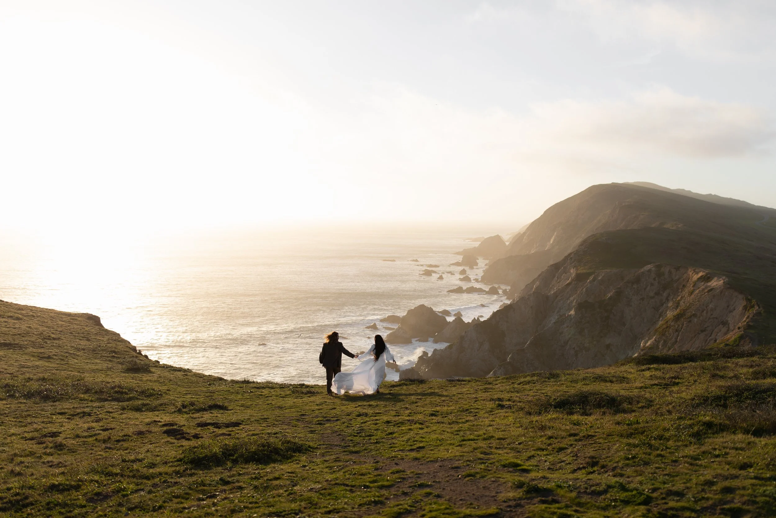 A couple holding hands on a grassy cliff overlooking the ocean at sunset, with rugged cliffs in the background.