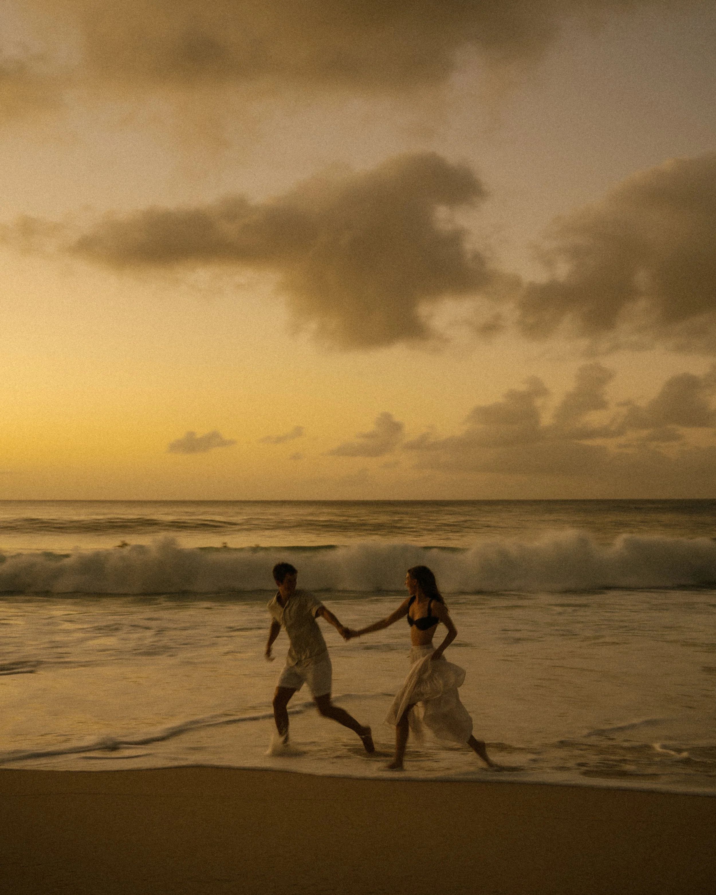 A couple holding hands while dancing on the beach at sunset with dark clouds overhead.