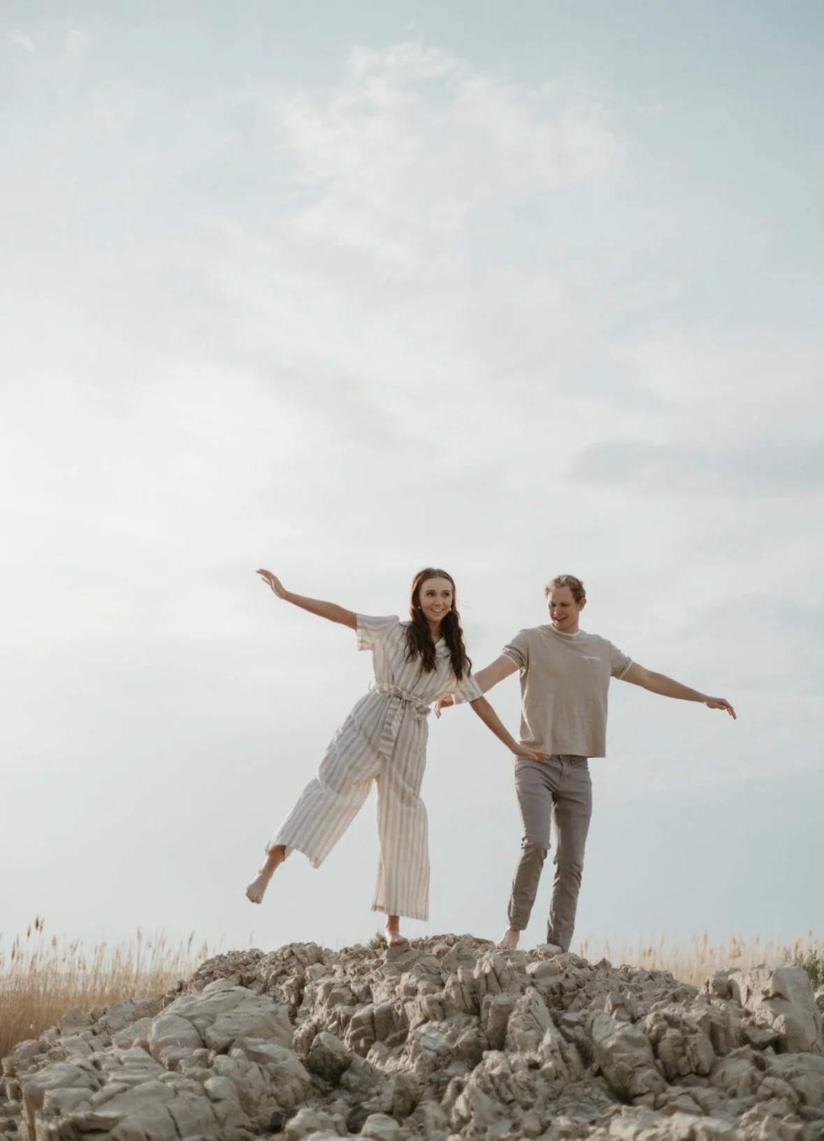 A young woman and man are balancing on a rocky surface outdoors, with the woman holding her arms out and the man stretching his arms out, under a cloudy sky.