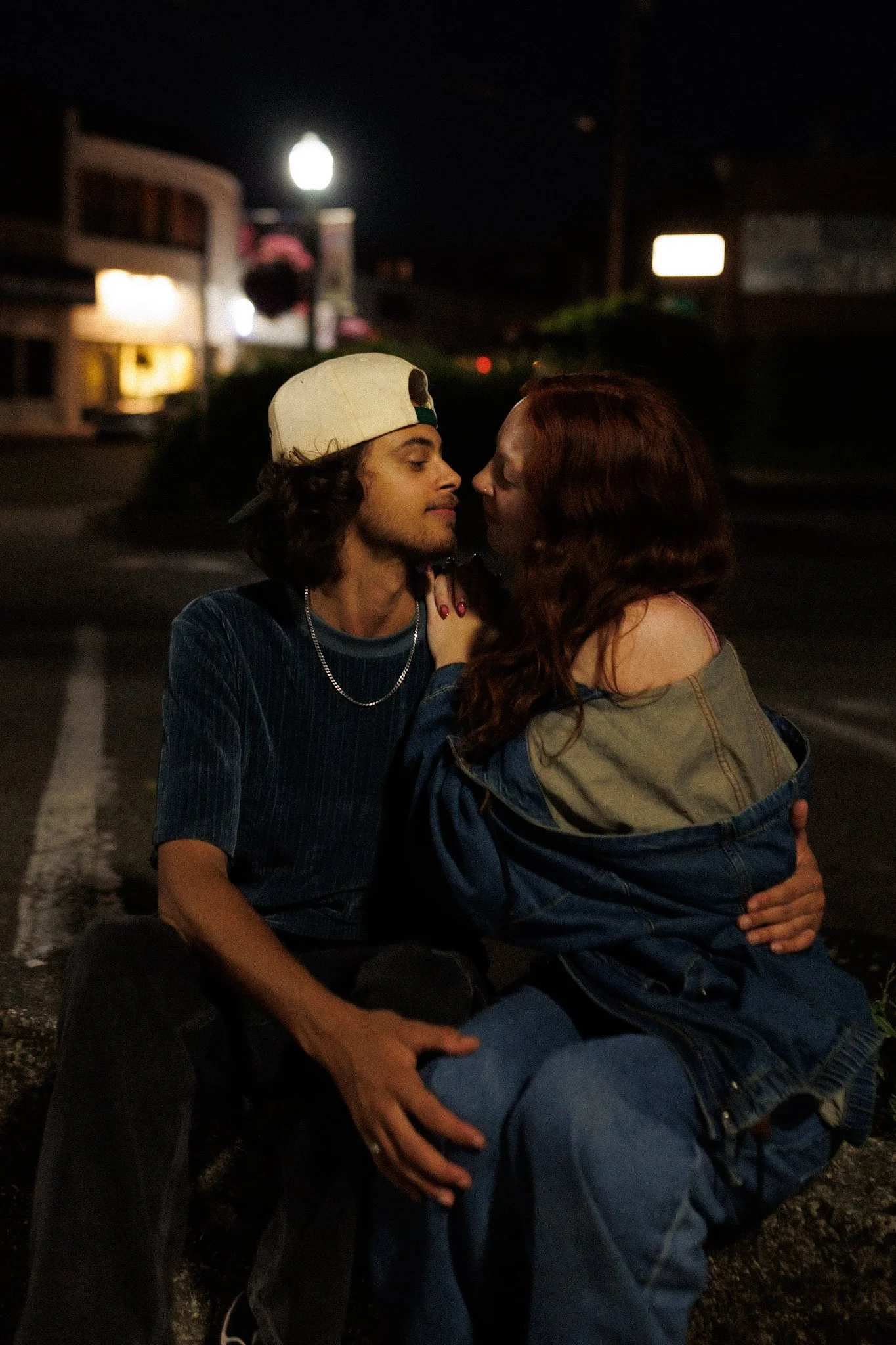 A young man and woman sit close together at night on the street, gazing into each other's eyes, with the woman holding the man's face gently.
