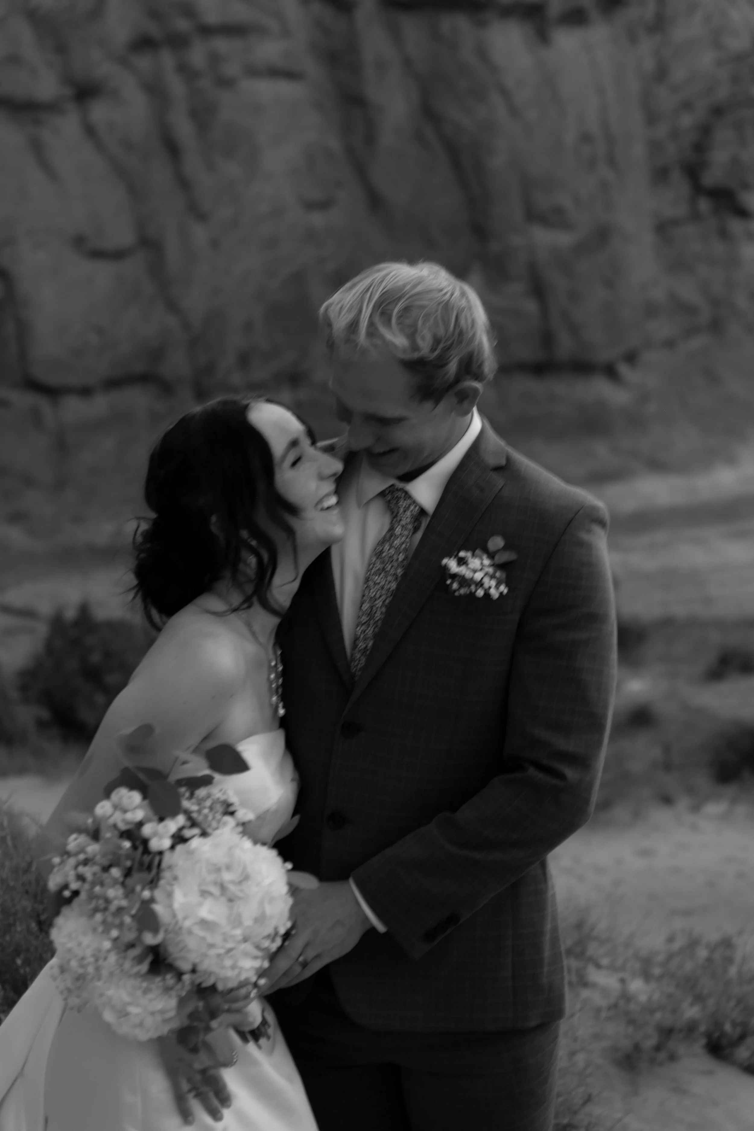 A black-and-white photo of a bride and groom sharing an intimate moment, the bride holding a bouquet of flowers, outdoors with rocky terrain in the background.