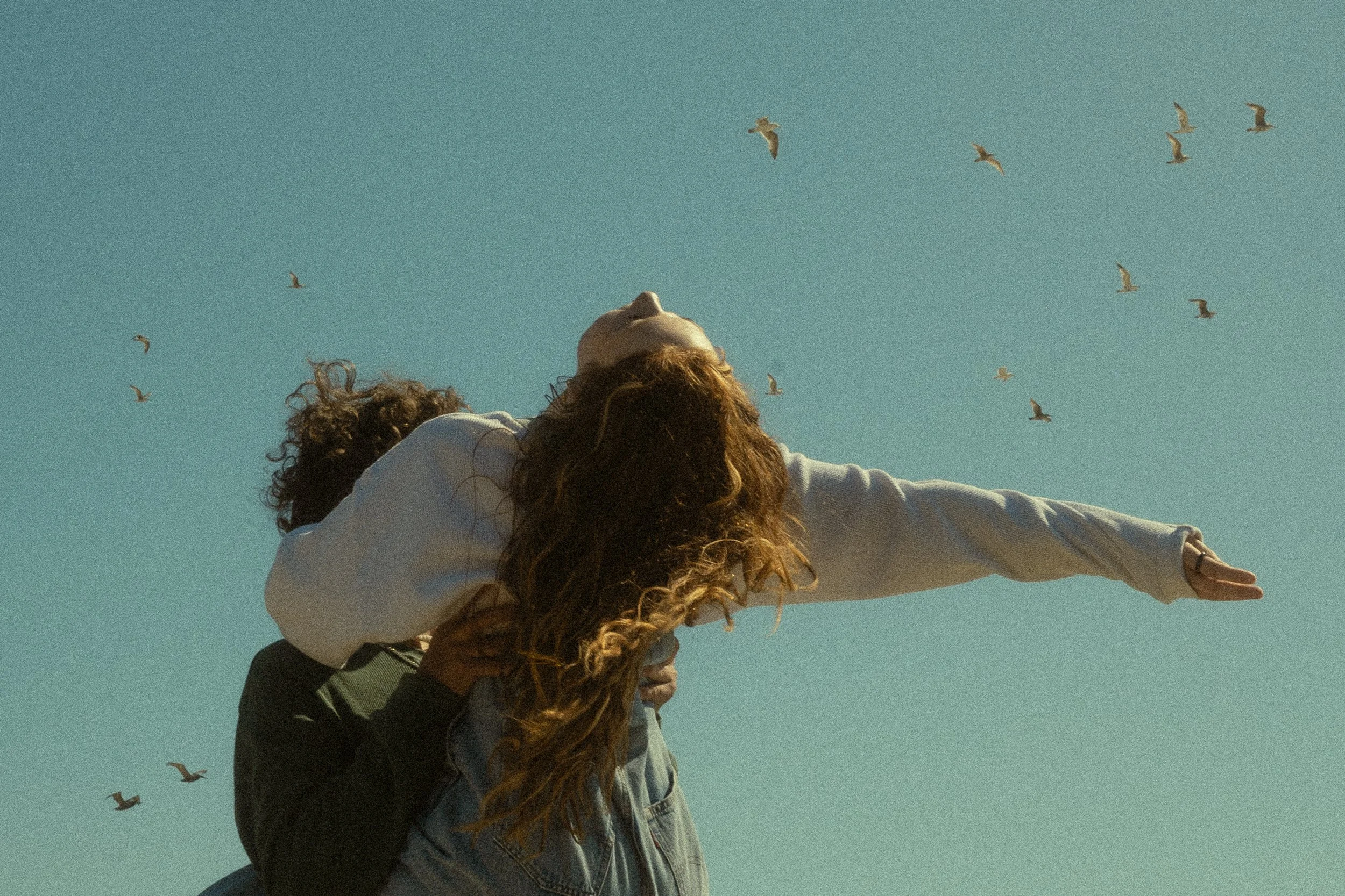 A woman with long curly hair, wearing a white hoodie, being carried on someone's back, extends her right arm outward in a stretch or gesture against a clear blue sky with seagulls flying overhead.