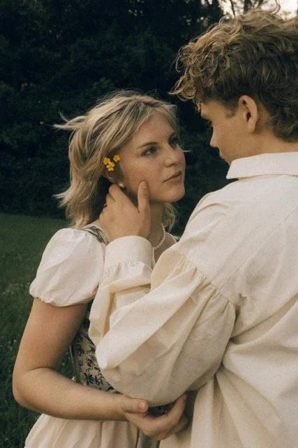 A young couple standing outdoors, facing each other, with the man gently touching the woman's face. The woman has blonde hair with a yellow flower accessory and is wearing a white dress, while the man has curly hair and is dressed in a light-colored shirt.