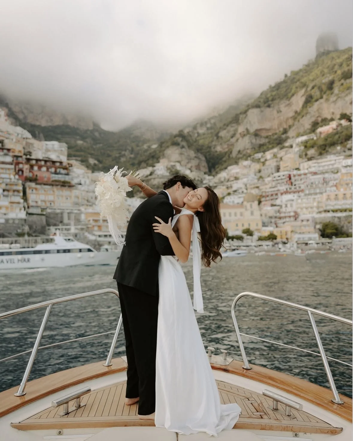 Happy couple in wedding attire sharing a kiss on a boat in a scenic harbor with hillside buildings and cloudy sky in the background.