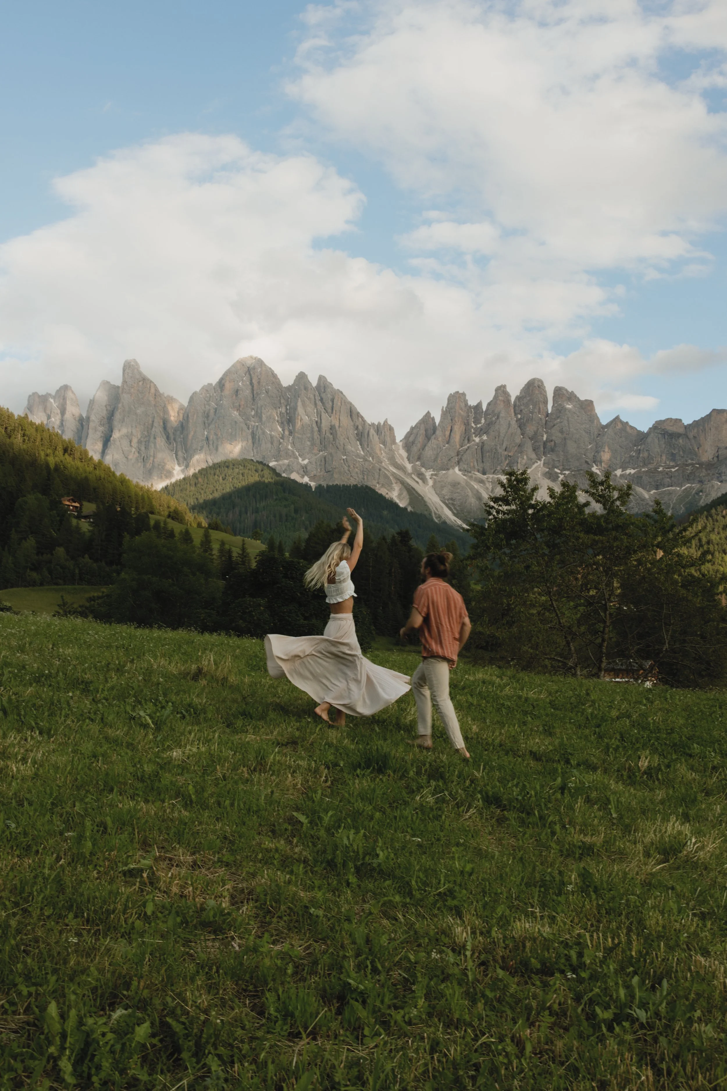 A couple dancing in a grassy field with mountains and trees in the background under a partly cloudy sky.
