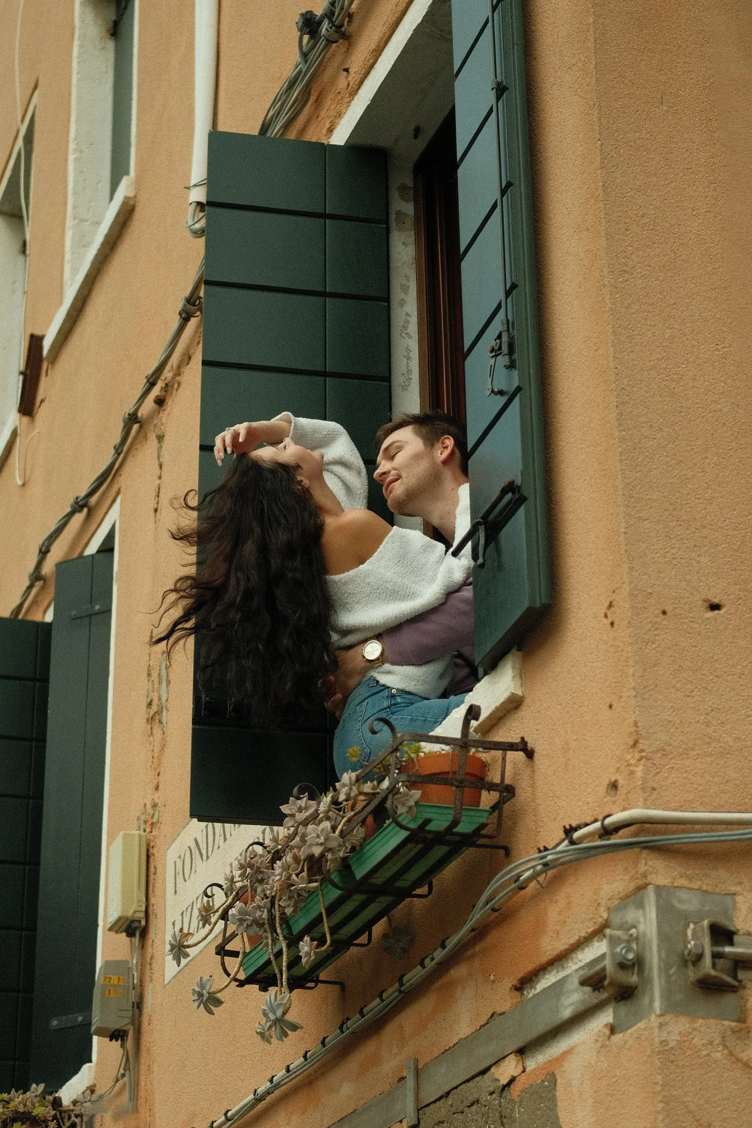 A couple embracing in a window with green shutters on an orange building.