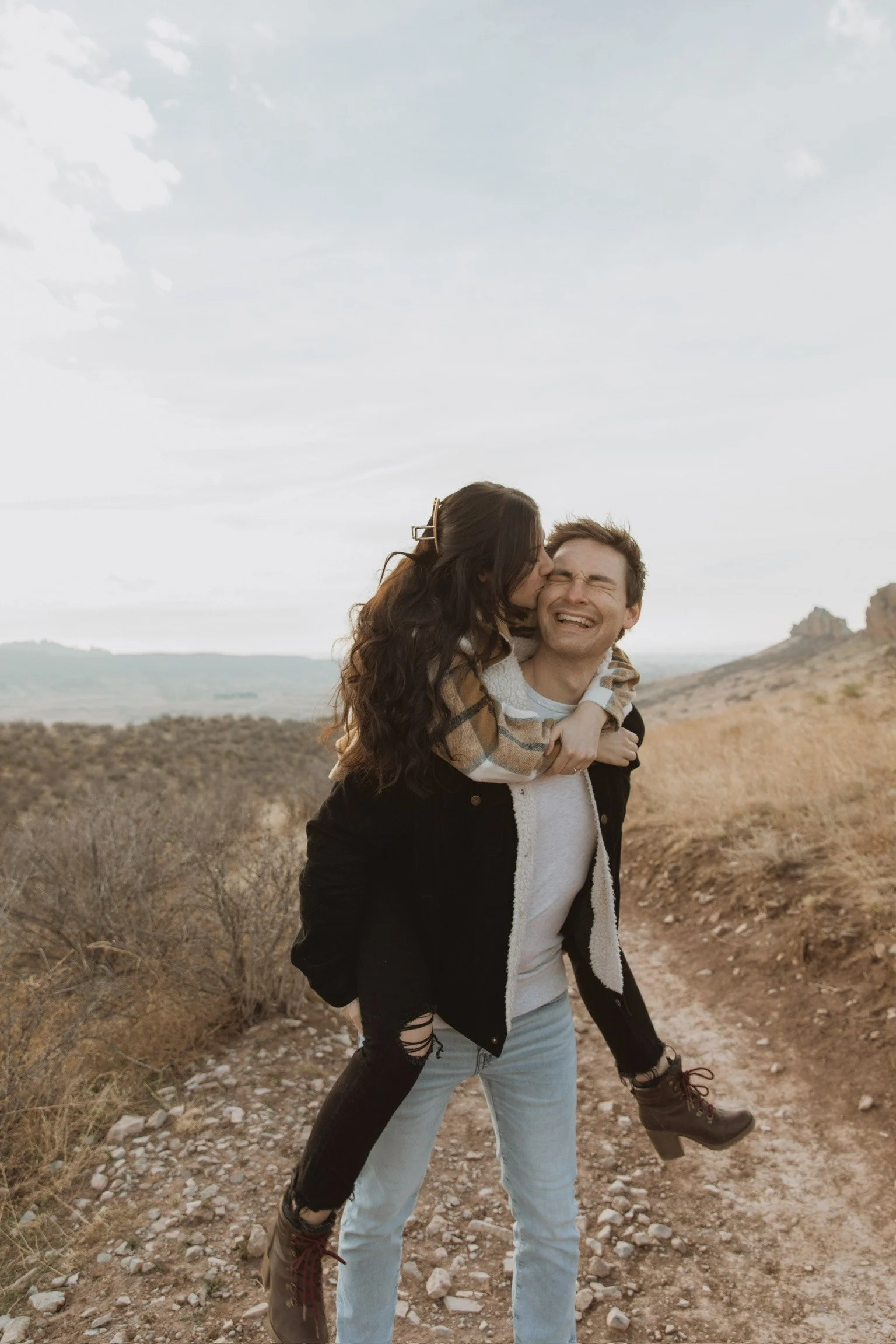 A young couple outdoors on a trail, with the woman giving the man a playful kiss on the cheek as he laughs, surrounded by dry grass and rolling hills in the background.