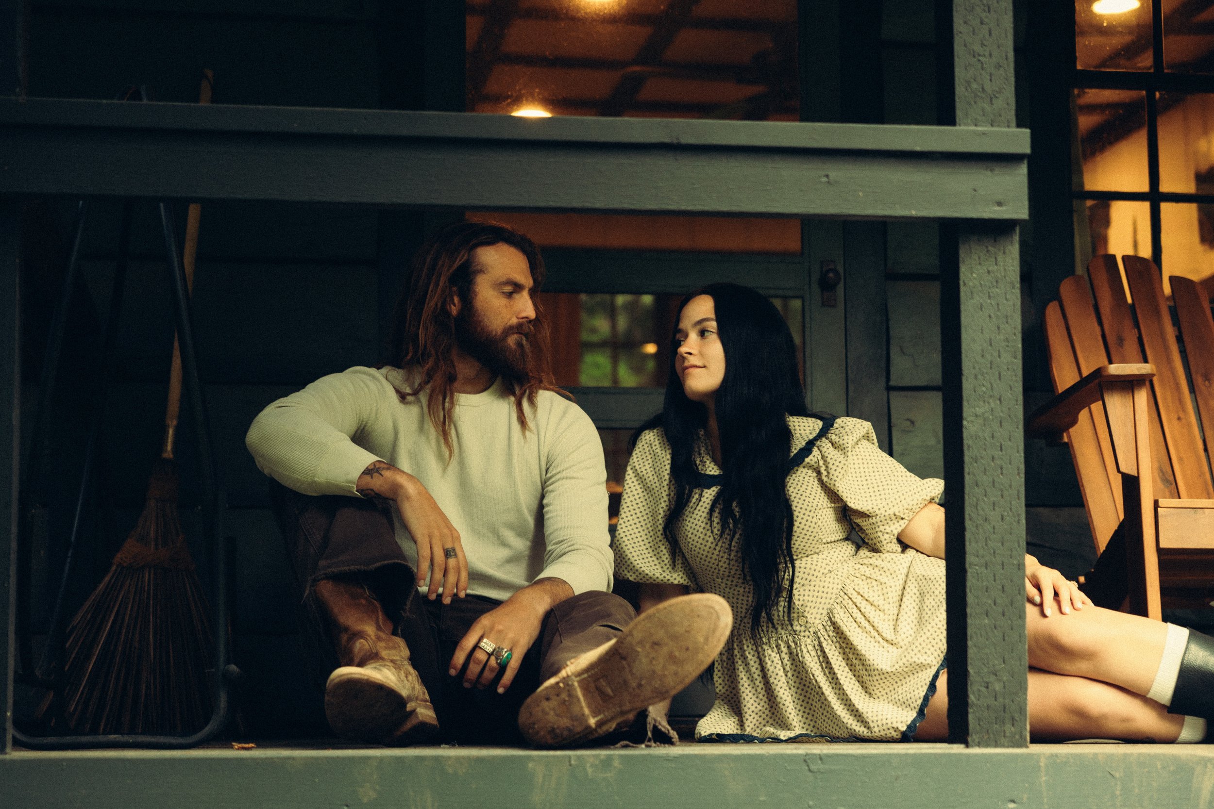 A man and woman sitting on the floor under a porch, looking at each other with a warm expression. The man has long hair, beard, and casual clothing, while the woman has long black hair, a yellow dress, and knee-high socks. There are wooden chairs and a broom nearby.