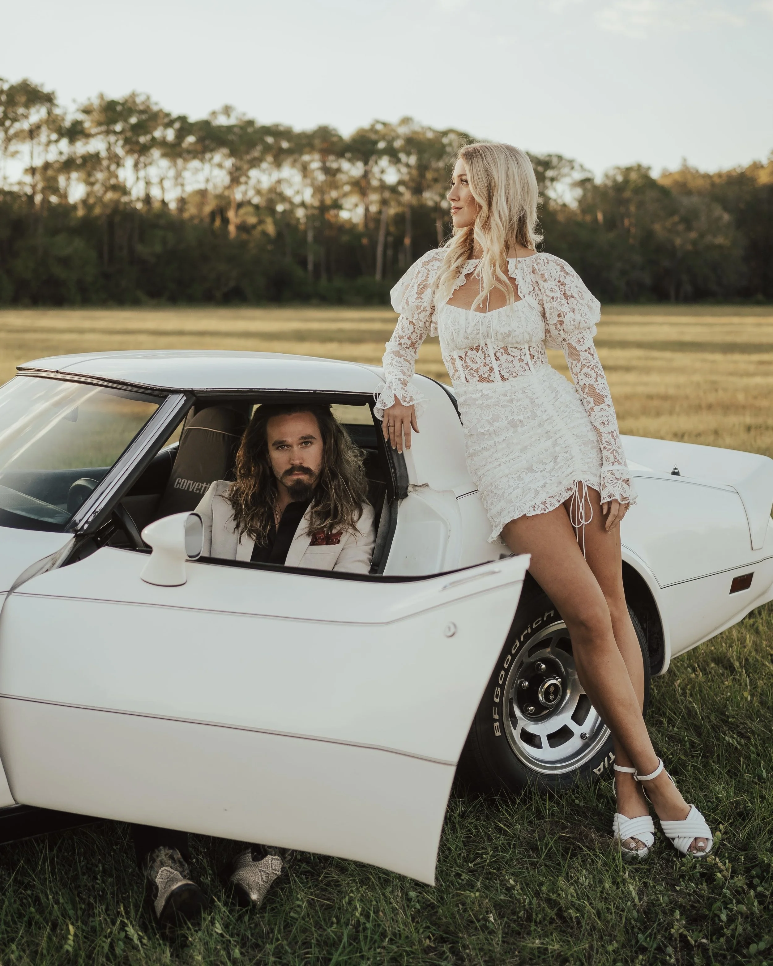 A man with long hair and a woman with blonde hair, both dressed in stylish clothing, posing with a white sports car outdoors during sunset. The woman is standing next to the car, leaning on the roof, while the man is sitting inside the car, looking out.