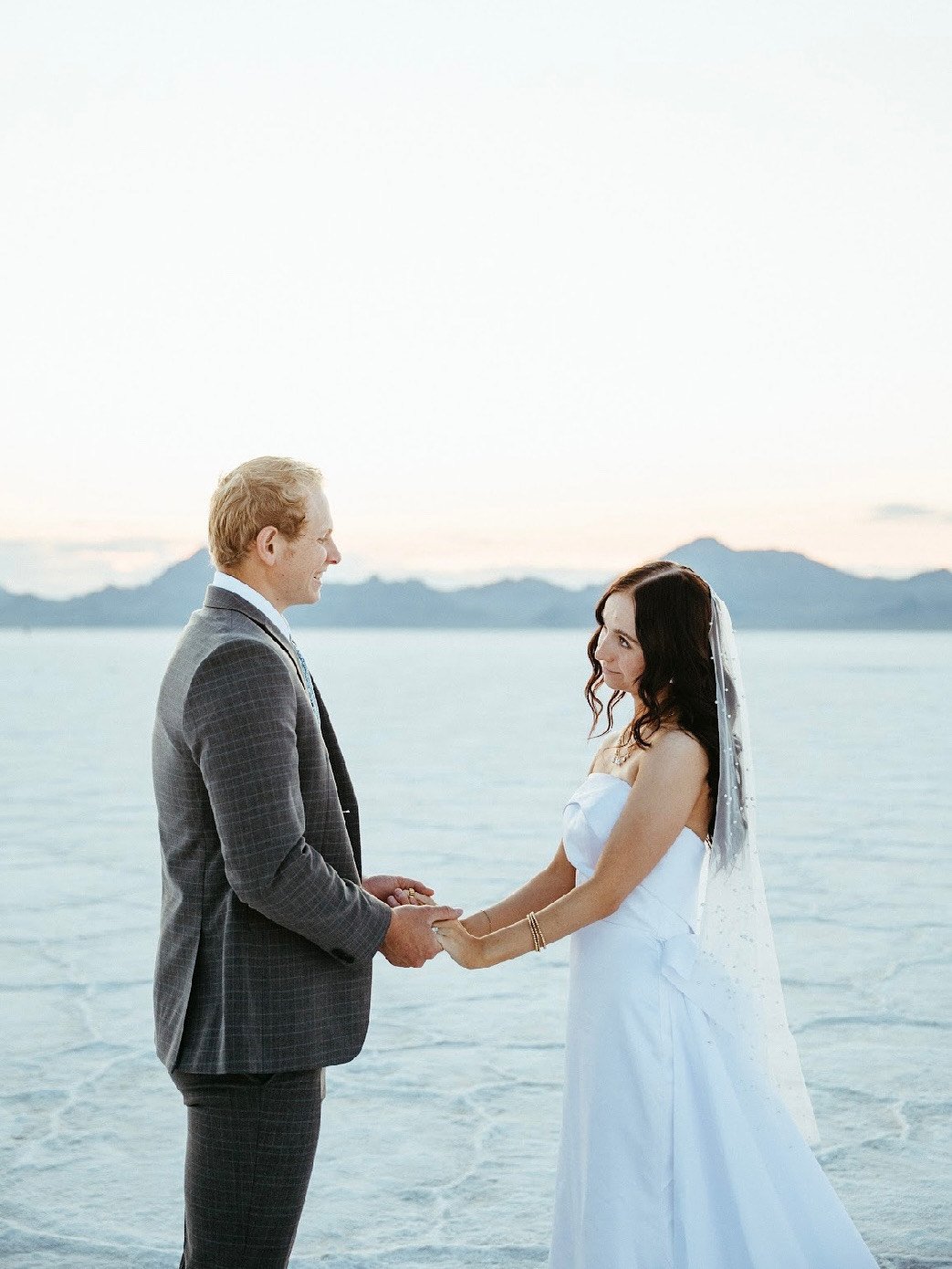 A bride and groom holding hands and facing each other on a beach at sunset, with mountains in the background, during their wedding ceremony.