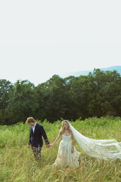 Bride and groom holding hands in a grassy field with trees in the background during daytime.