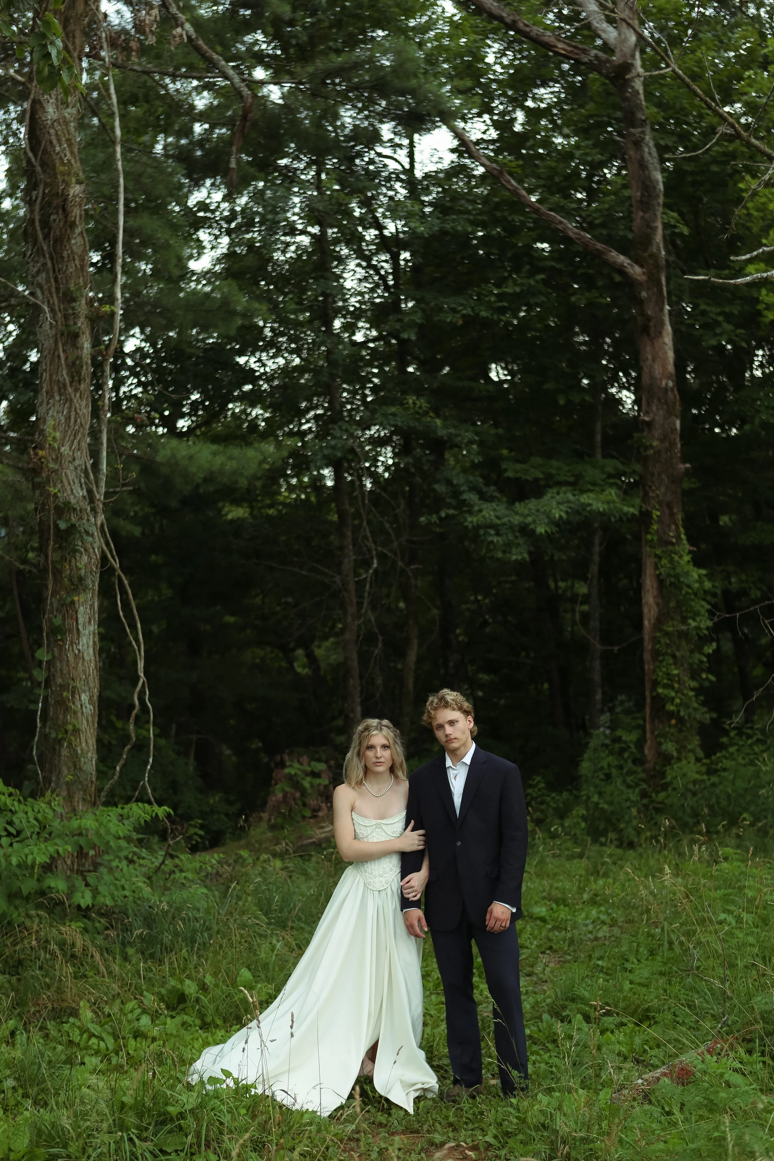A couple dressed in formal attire, with the woman in a white gown and the man in a black suit, standing in a lush green forest.