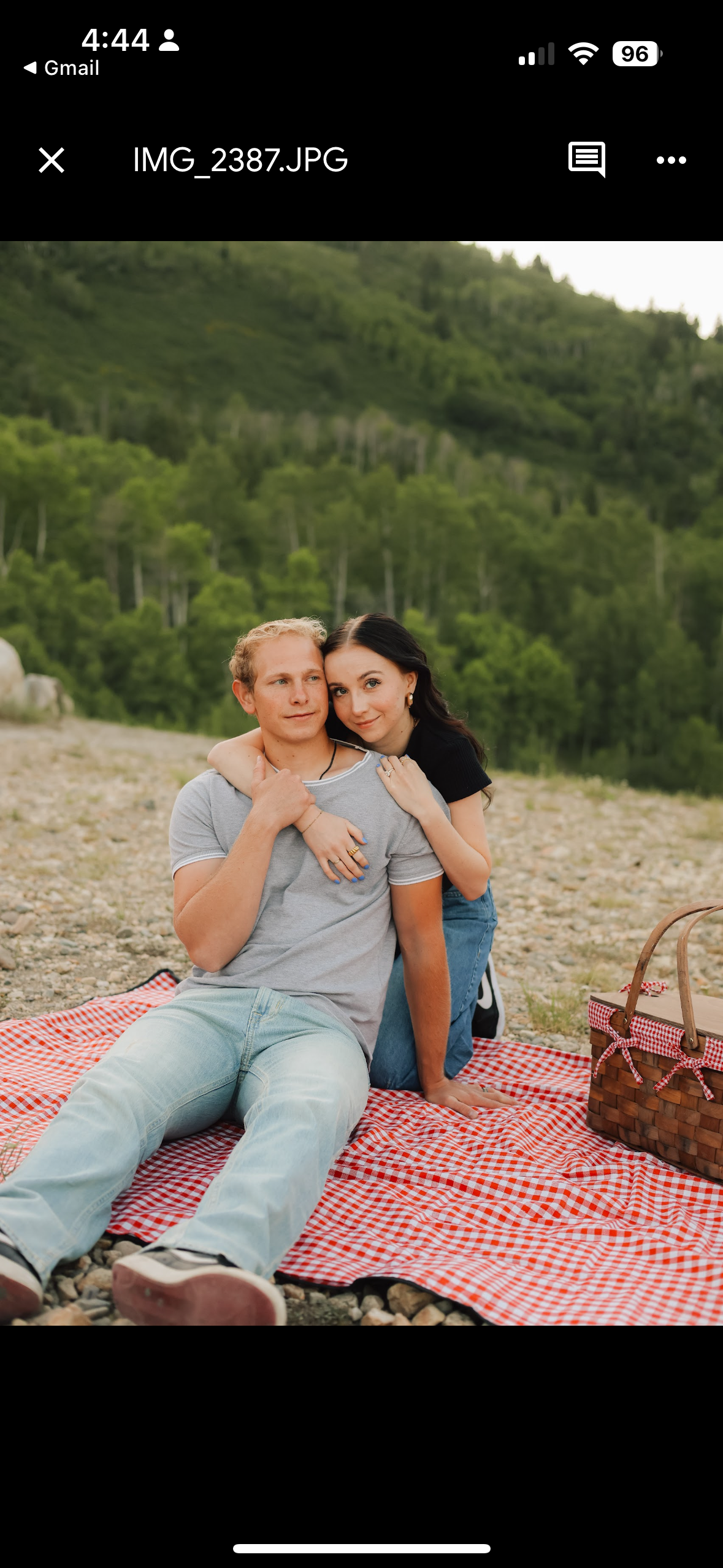 A young couple sitting on a red and white checkered picnic blanket outdoors, with a lush green forested hillside in the background. The woman is hugging the man from behind, resting her head on his, while the man sits with his legs extended and looks to the side. There is a picnic basket on the blanket.