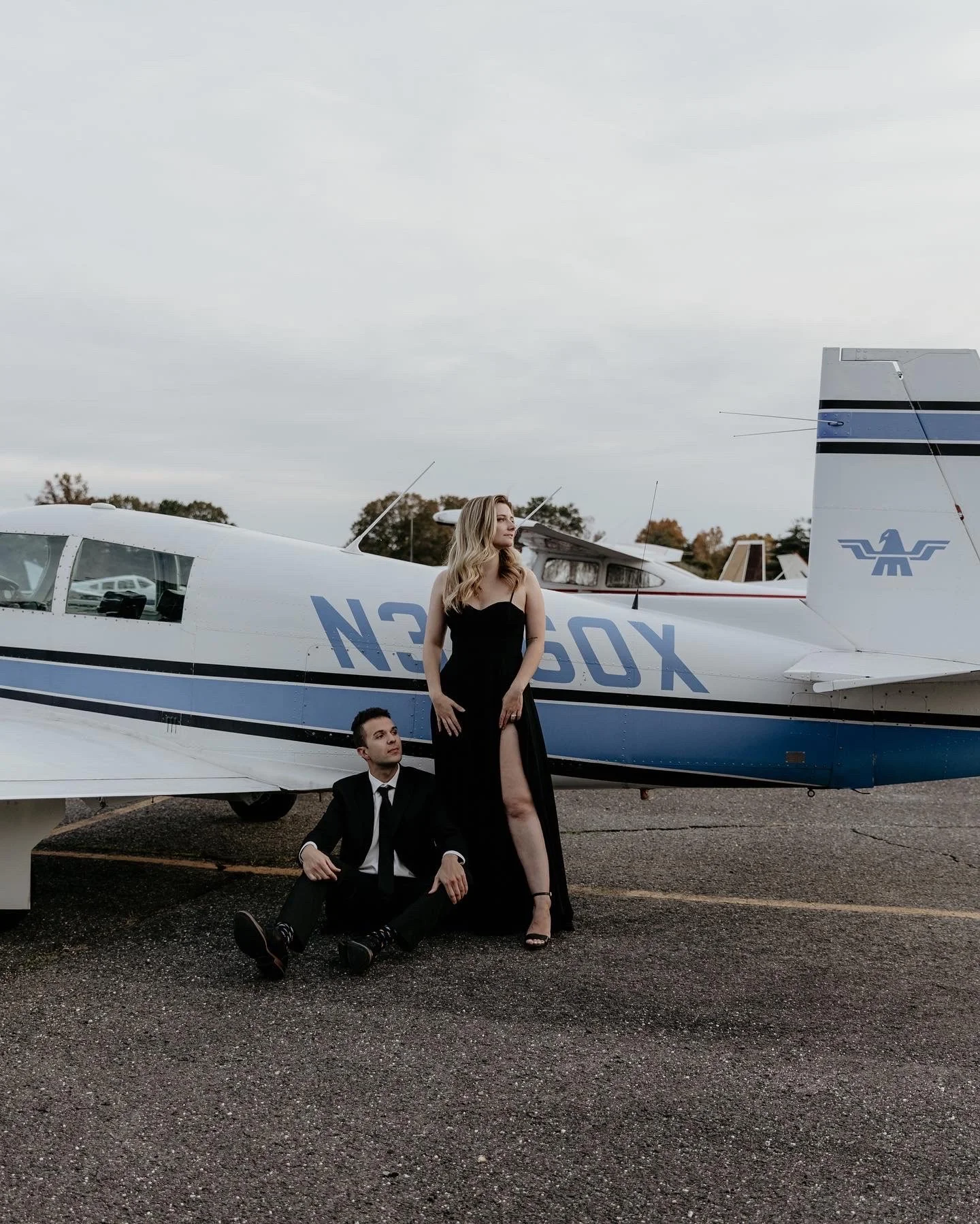 A woman in a black evening gown with a high slit standing next to a man in a black suit, sitting on the ground, in front of a small propeller airplane at an airfield on a cloudy day.