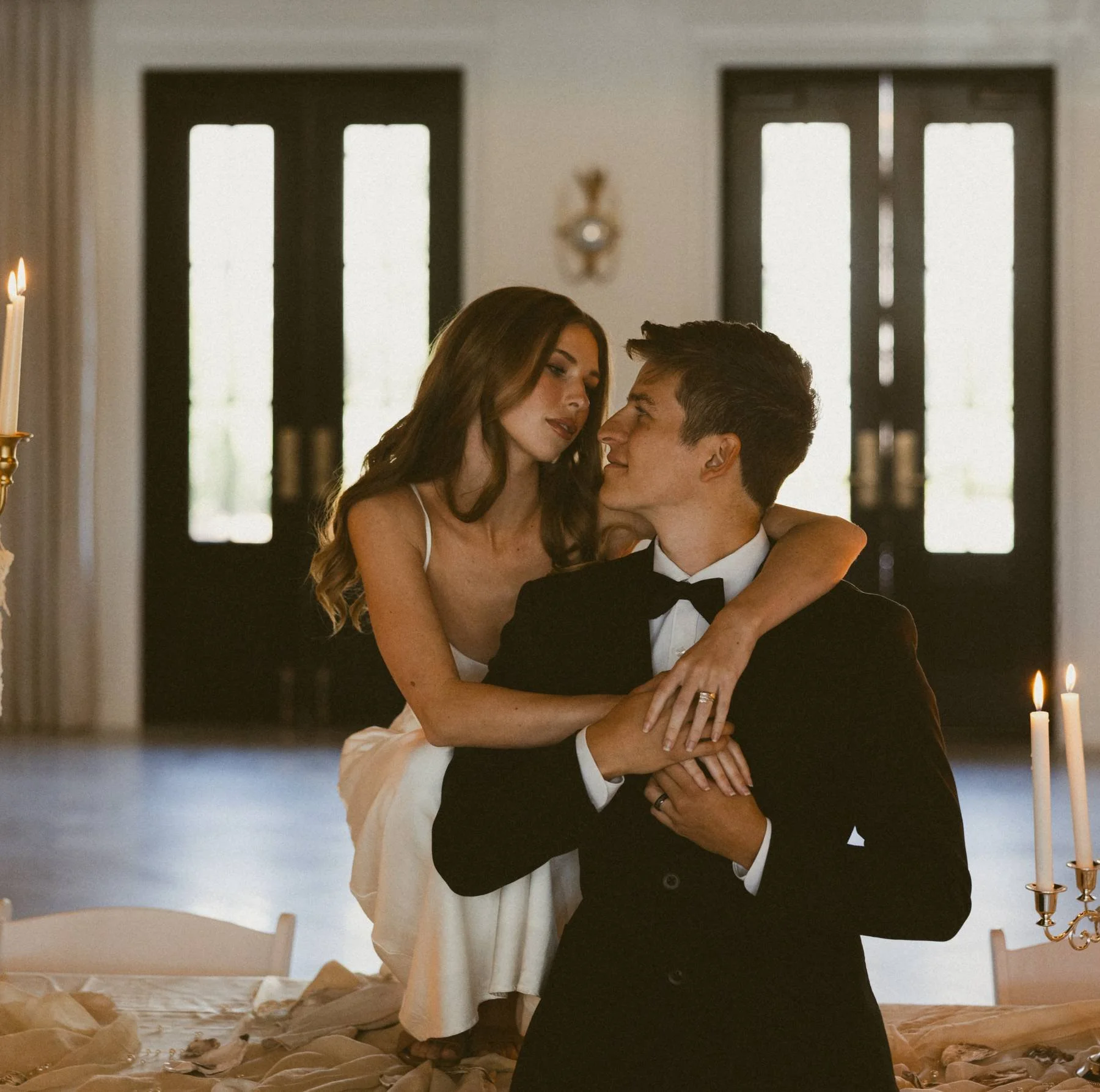 A couple dressed in wedding attire, with the woman sitting on a table and the man standing, gazing at each other in a warmly lit room with candles and double doors in the background.