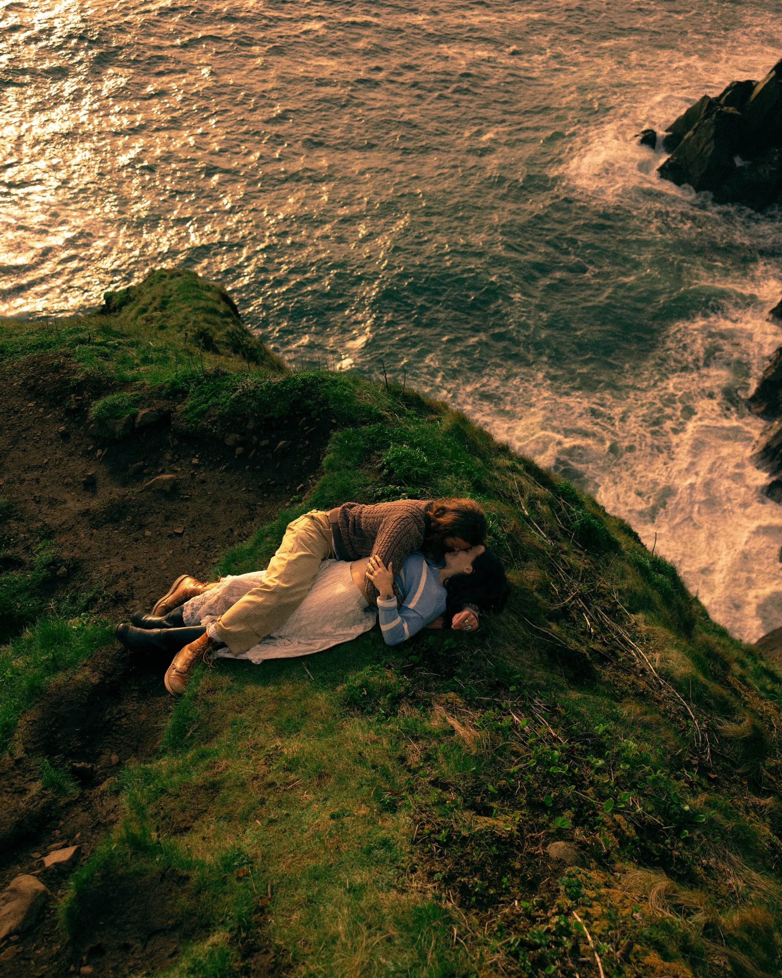 Two people lying on a grassy cliffside near the ocean, with sunset lighting casting a warm glow.