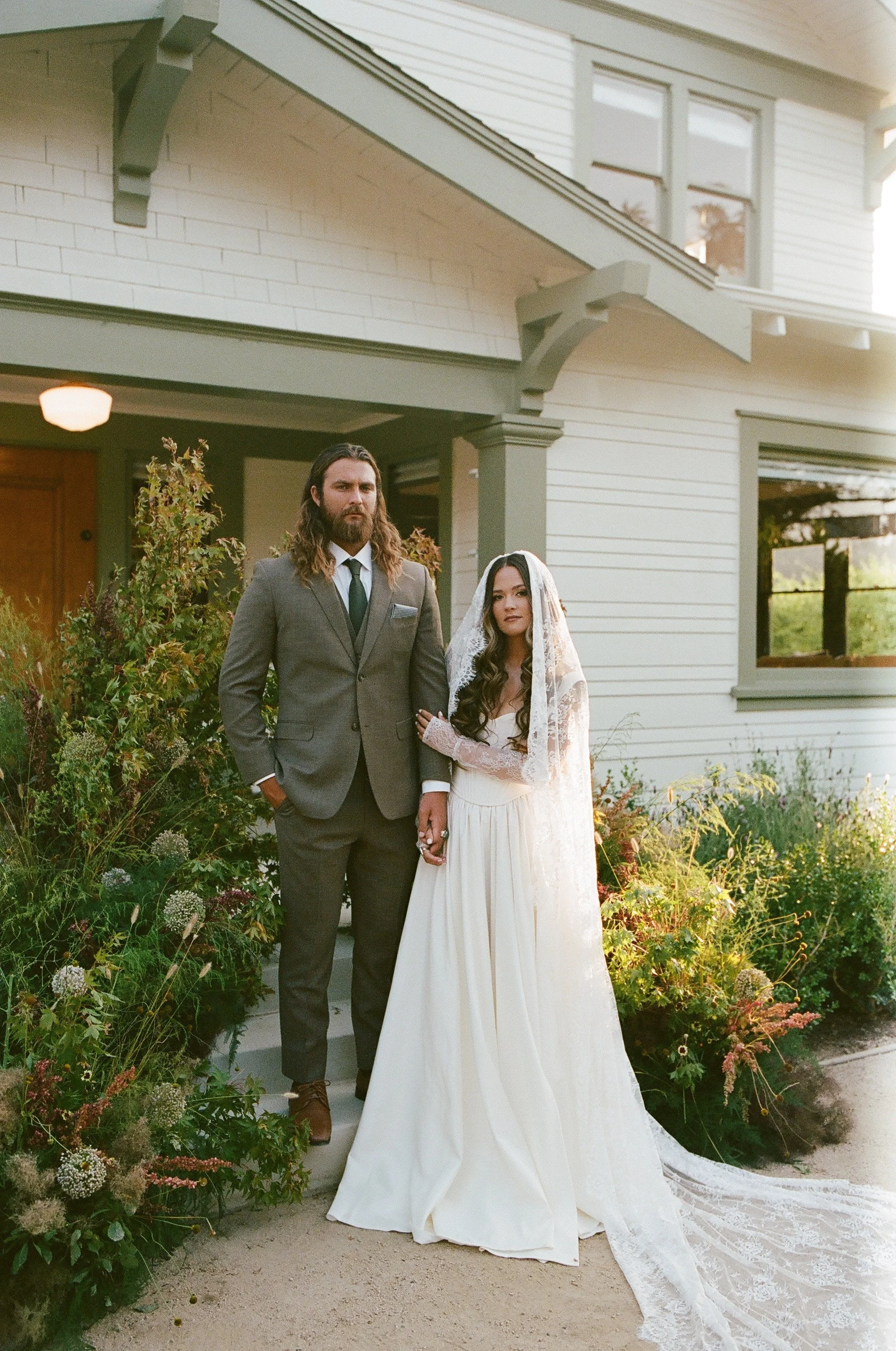 A bride and groom stand outside a house, holding hands, with the bride wearing a white wedding dress and veil, and the groom in a gray suit, with greenery and flowers surrounding them during sunset.