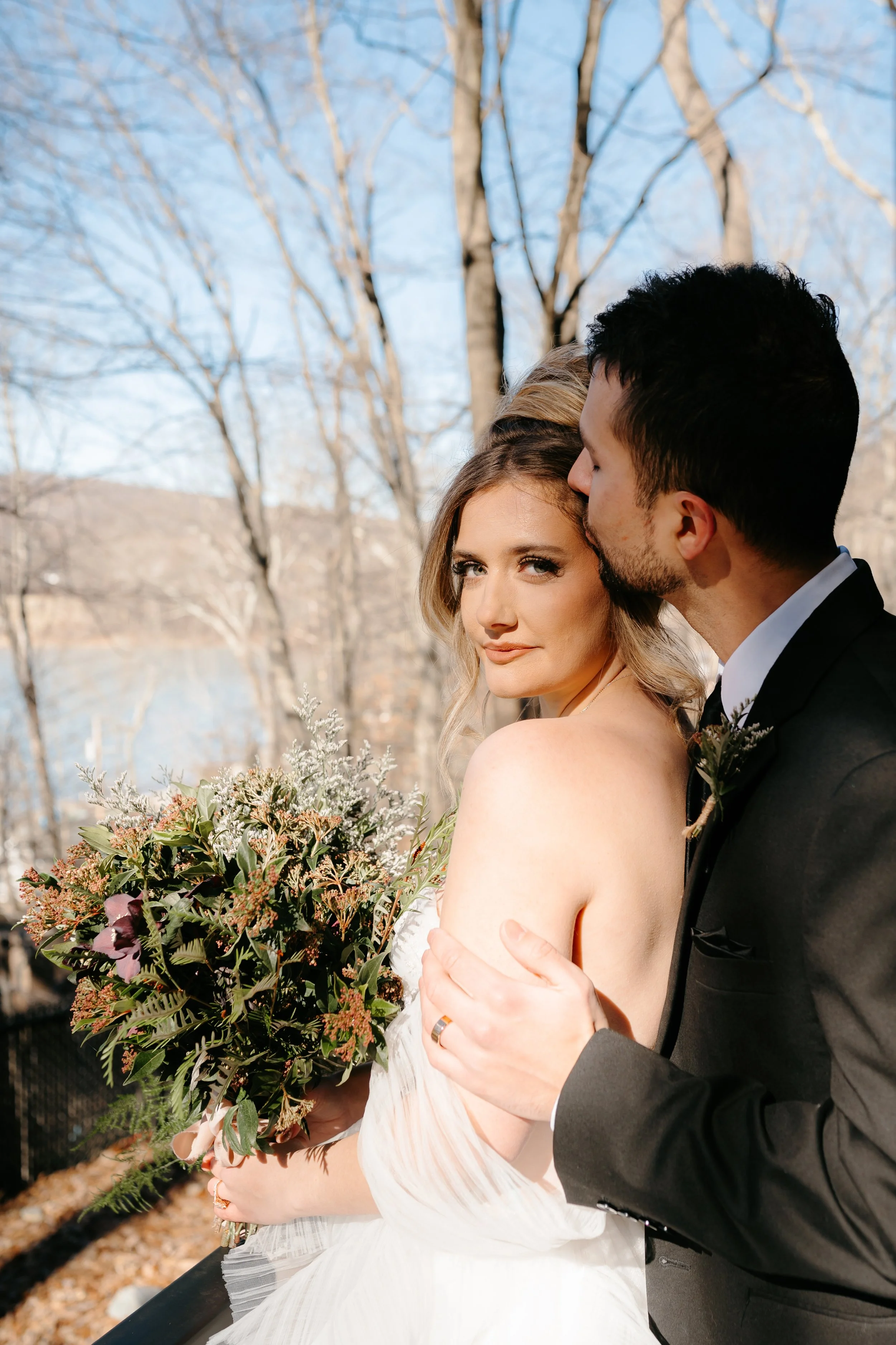 A newly married couple standing outdoors, with the groom kissing the bride's temple. The bride holds a bouquet of flowers and looks at the camera, while trees and a lake are in the background on a clear day.