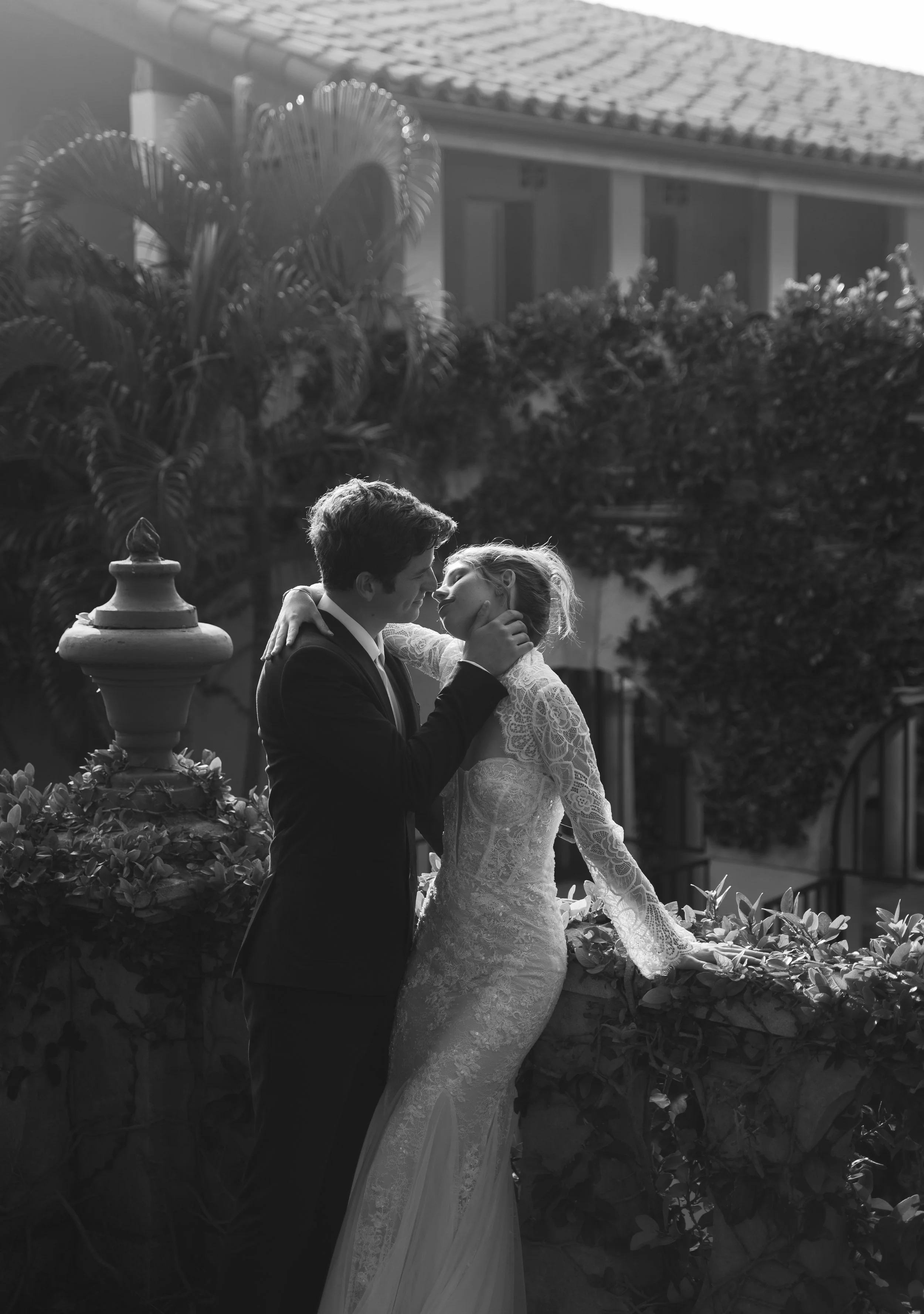 A black and white photo of a couple on their wedding day, with the man holding the woman's face and leaning in for a kiss, standing outdoors near a garden wall.