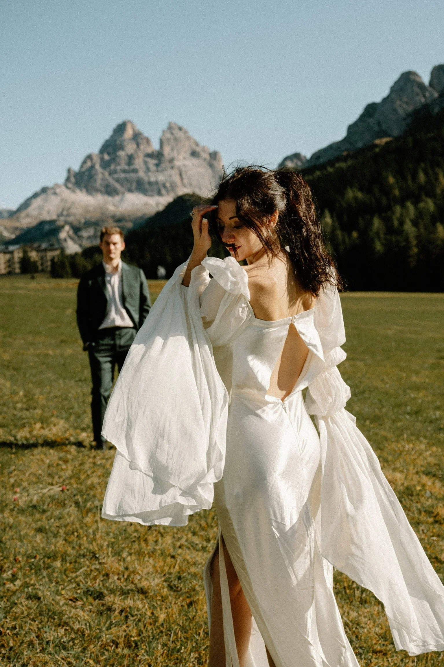 A woman in a white dress standing in a grassy field with mountains in the background, a man in a suit approaching her.