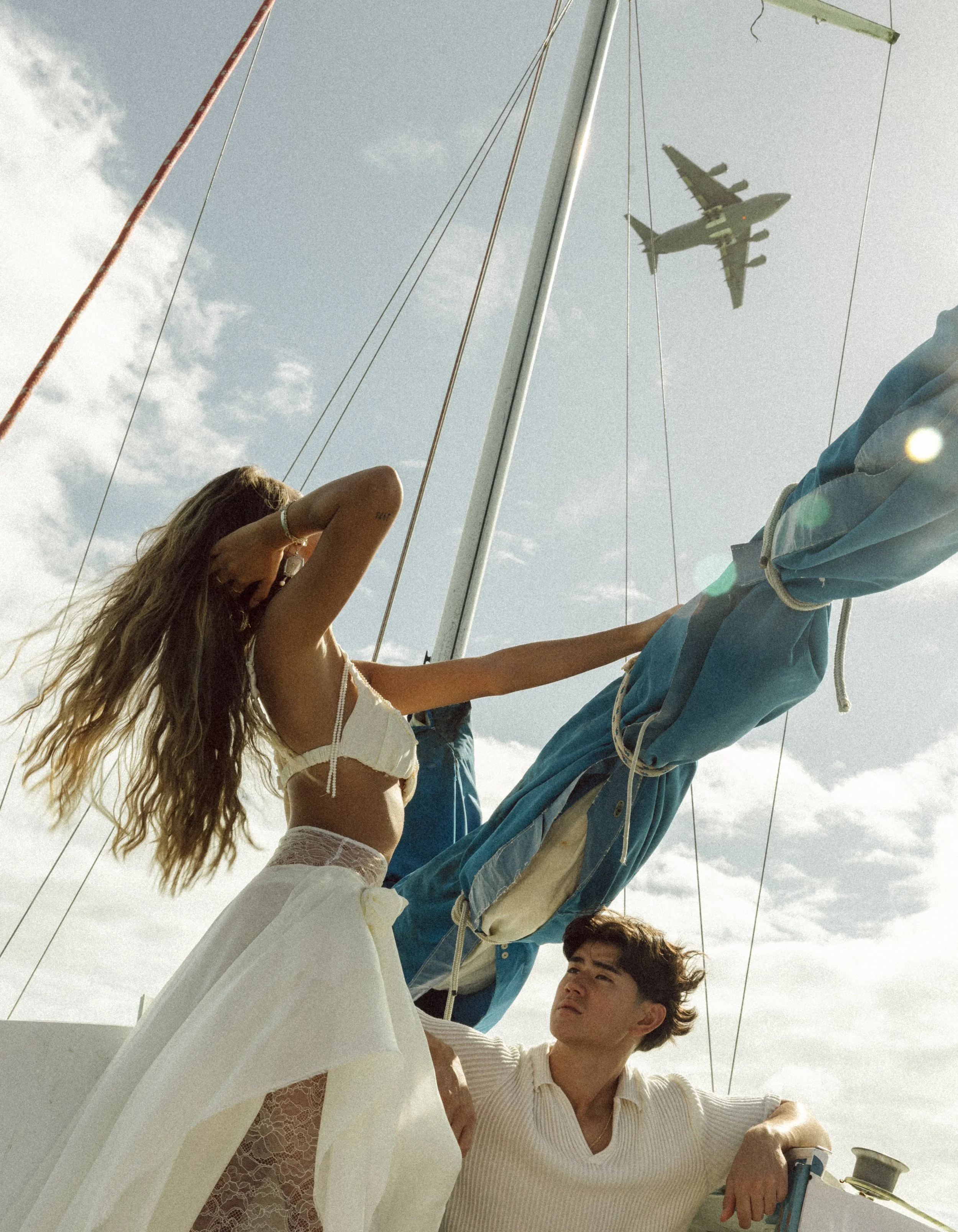 A young couple on a sailboat, with the woman adjusting the sail and the man sitting nearby, under a partly cloudy sky with an airplane flying overhead.