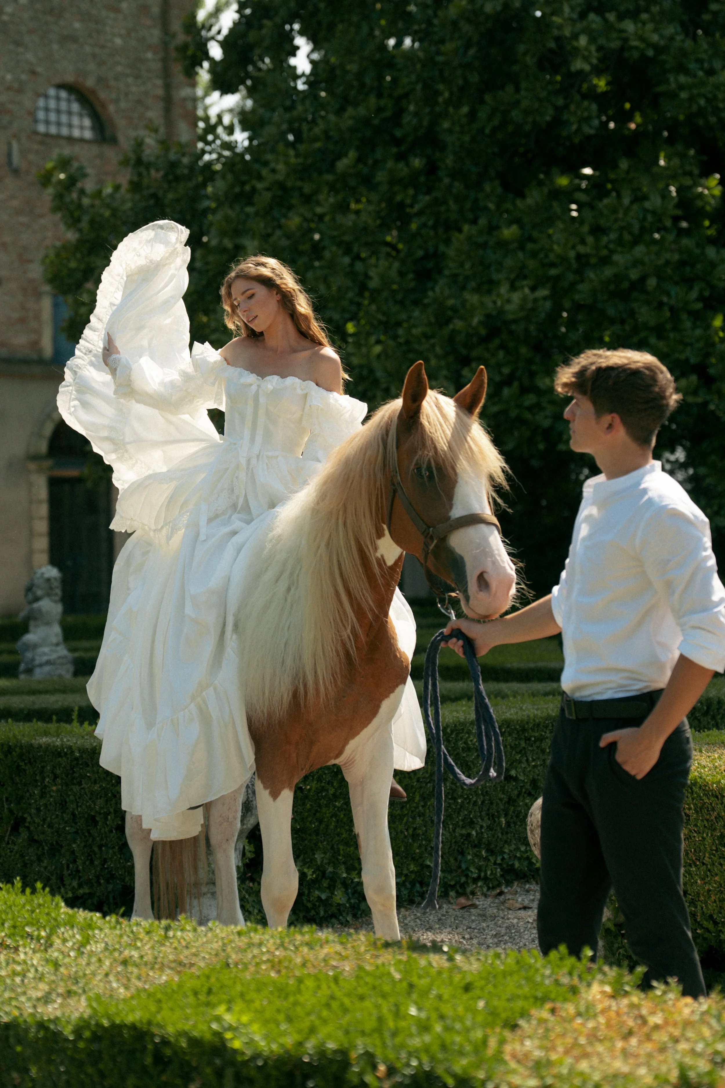 A model in a white wedding dress posing on a horse, talking to a young man with brown hair holding the horse's bridle outside in a garden.