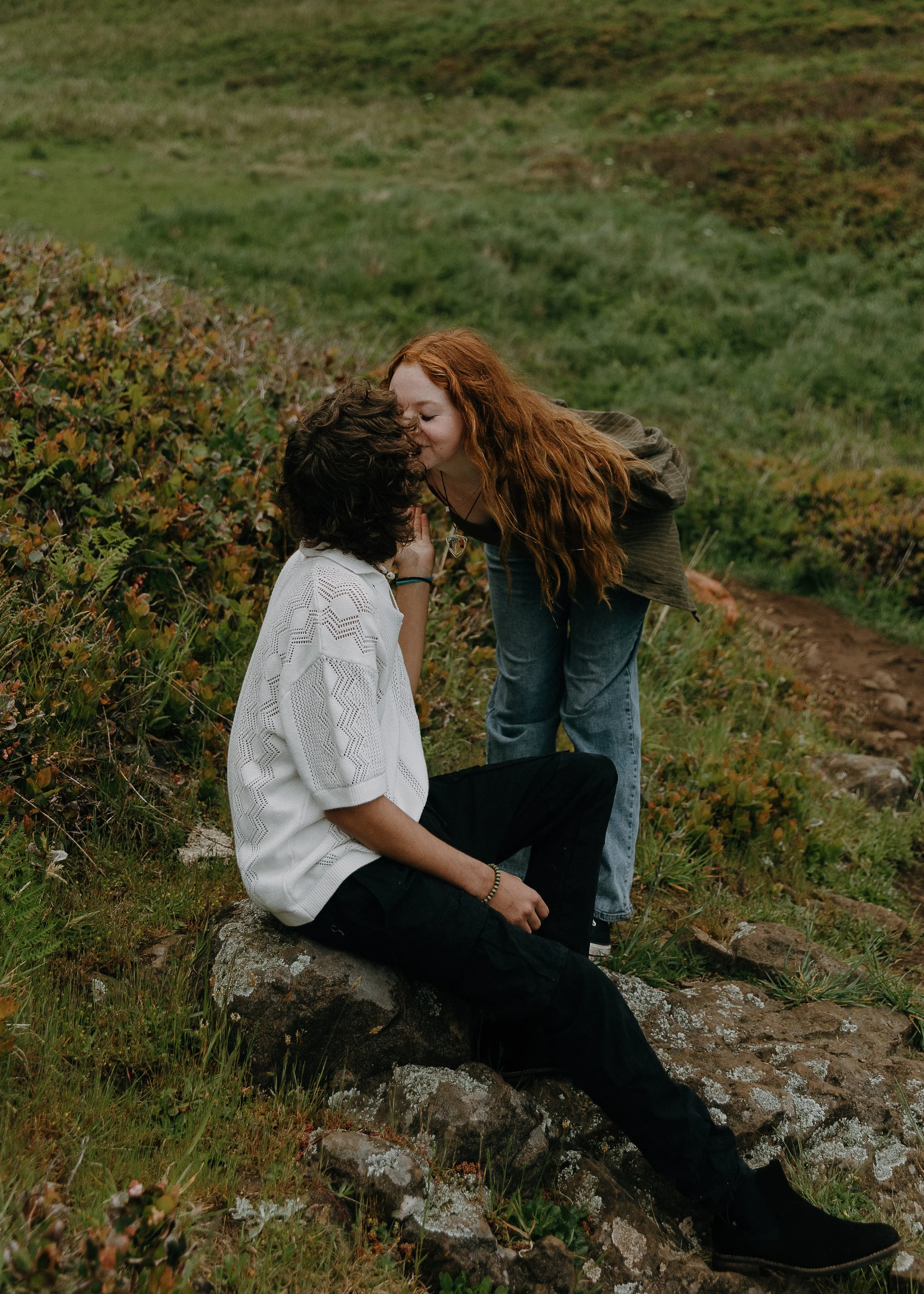 A couple sharing a kiss outdoors on a grassy hillside with mountains in the background, during what appears to be late afternoon or early evening.