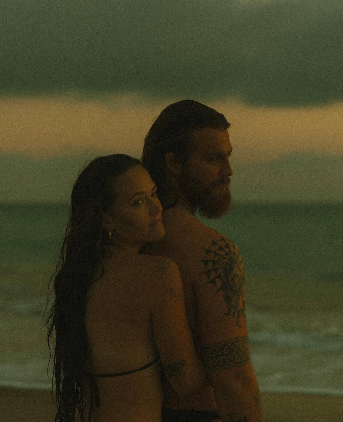 A man and woman with tattoos standing on the beach at dusk, facing sideways with the ocean and waves in the background.