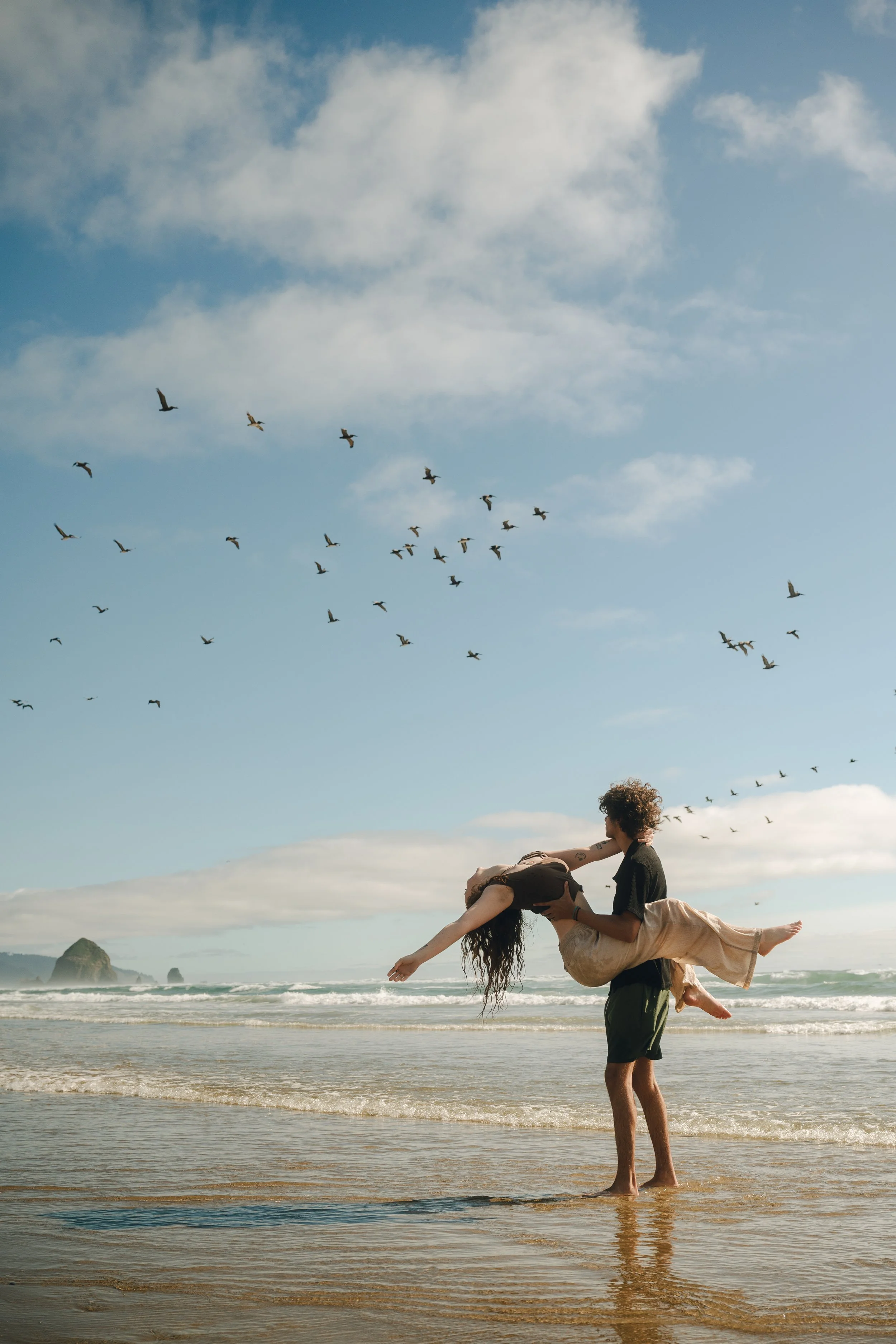 A couple on the beach, with the man holding the woman in his arms as she leans back, near the shoreline with seagulls flying overhead and rock formations in the distance under a partly cloudy sky.